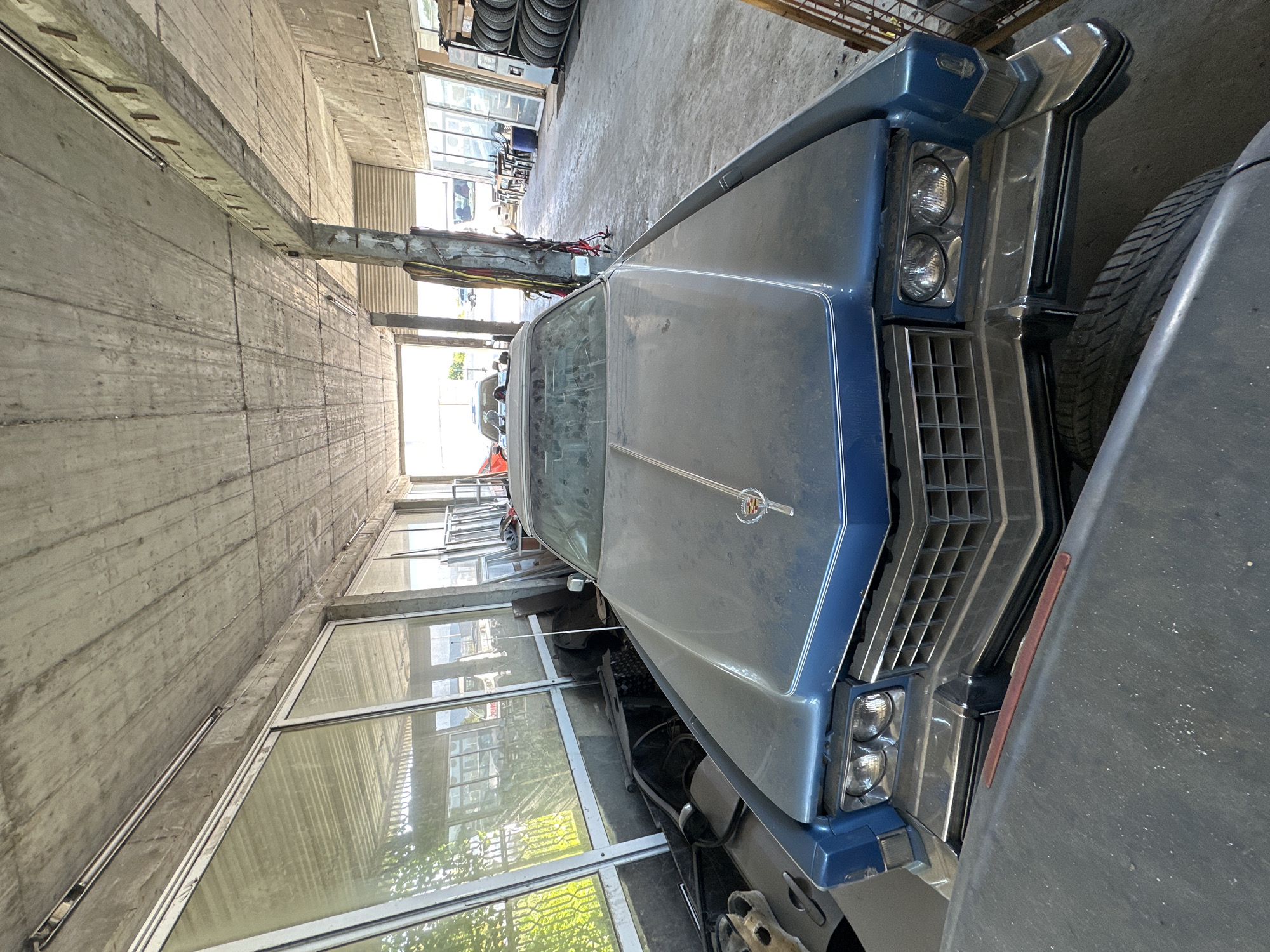 A blue Cadillac is parked inside a garage. The car is covered in dust. The garage has a concrete ceiling and columns, and large windows on one side. Other vehicles and tires are visible in the background.