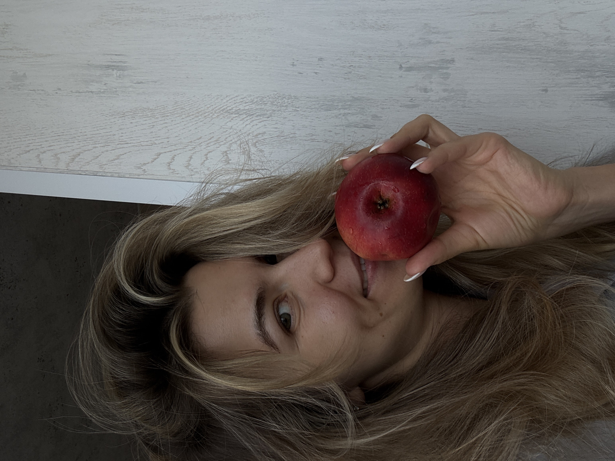 A person is depicted in a portrait-style image, rotated 90 degrees clockwise. The main subject is a woman with long, wavy blonde and light brown hair, which is spread out around her head. She is holding a red apple with her right hand, positioning it near her face to partially obscure one eye. Her visible eye is looking towards the viewer, and she has a slight, friendly smile on her lips, revealing some teeth.
Her hand has white-tipped manicured nails (French manicure). The apple appears fresh and has tiny water droplets on its surface.
The background consists of two distinct areas: a light-colored, possibly white or pale gray, wooden or laminate surface with a visible wood grain texture, occupying the upper portion of the rotated image. Below this, there is a darker, mottled grey or black surface, possibly a wall or floor.
There is no visible text in the image. The setting appears to be indoors, but no specific location cues are present beyond the generic surfaces. The lighting suggests an indoor environment.
