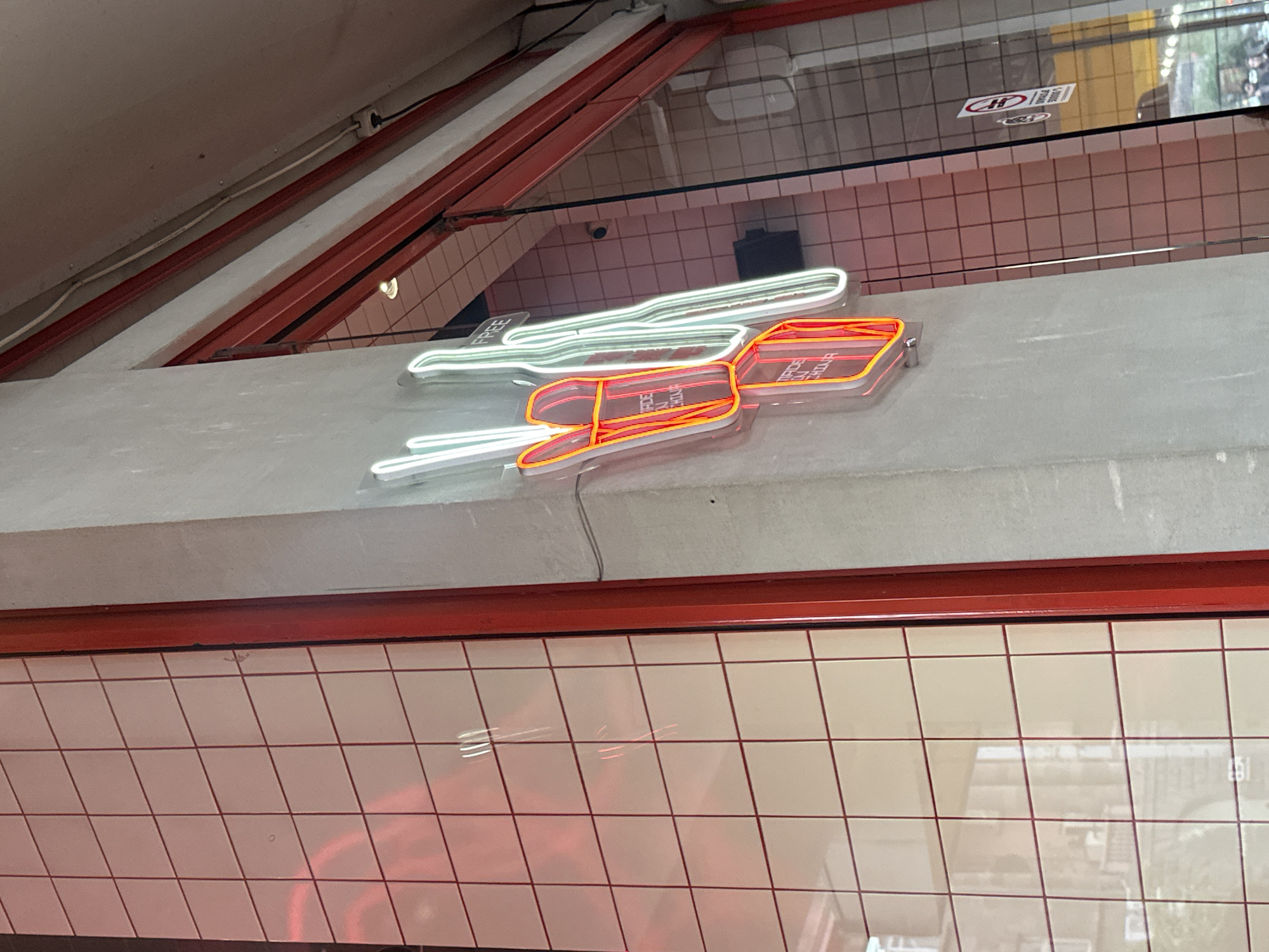 The image shows a neon sign that reads "Made in China," located on a gray ledge above a tiled wall. The sign is white and red, with the words arranged in stacked rectangles. The wall is covered in square tiles with reddish grout. The sign is illuminated. The location in the city of Yerevan, Armenia cannot be determined.