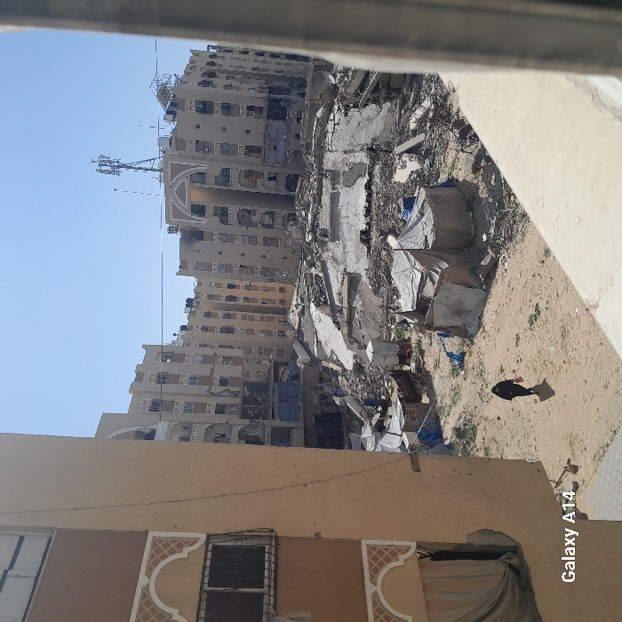The image displays an extensively damaged urban area in Khan Yunis, Palestinian Territory, under a clear blue sky. The perspective is from an elevated vantage point, with the scene framed on the left by a partial view of a light-colored building exhibiting a barred window and decorative facade elements. On the right, a concrete railing partially obstructs the view.
The midground features a broad expanse of sandy, disturbed earth, heavily strewn with structural debris, including large concrete slabs and fragmented building materials, indicative of significant demolition. Scattered within this area are multiple makeshift shelters constructed from tarpaulins and various fabrics, some featuring small satellite dishes. A single individual, clad in dark clothing, walks from left to right across the sandy terrain, casting a distinct shadow and carrying a light-colored object in their right hand.
In the background, a row of multi-story residential buildings stands, all exhibiting widespread and severe damage. Numerous windows are broken or missing, building facades are extensively pockmarked and perforated, and sections of balconies and walls appear collapsed or destroyed. A prominent telecommunications tower is visible atop one of these damaged background structures.