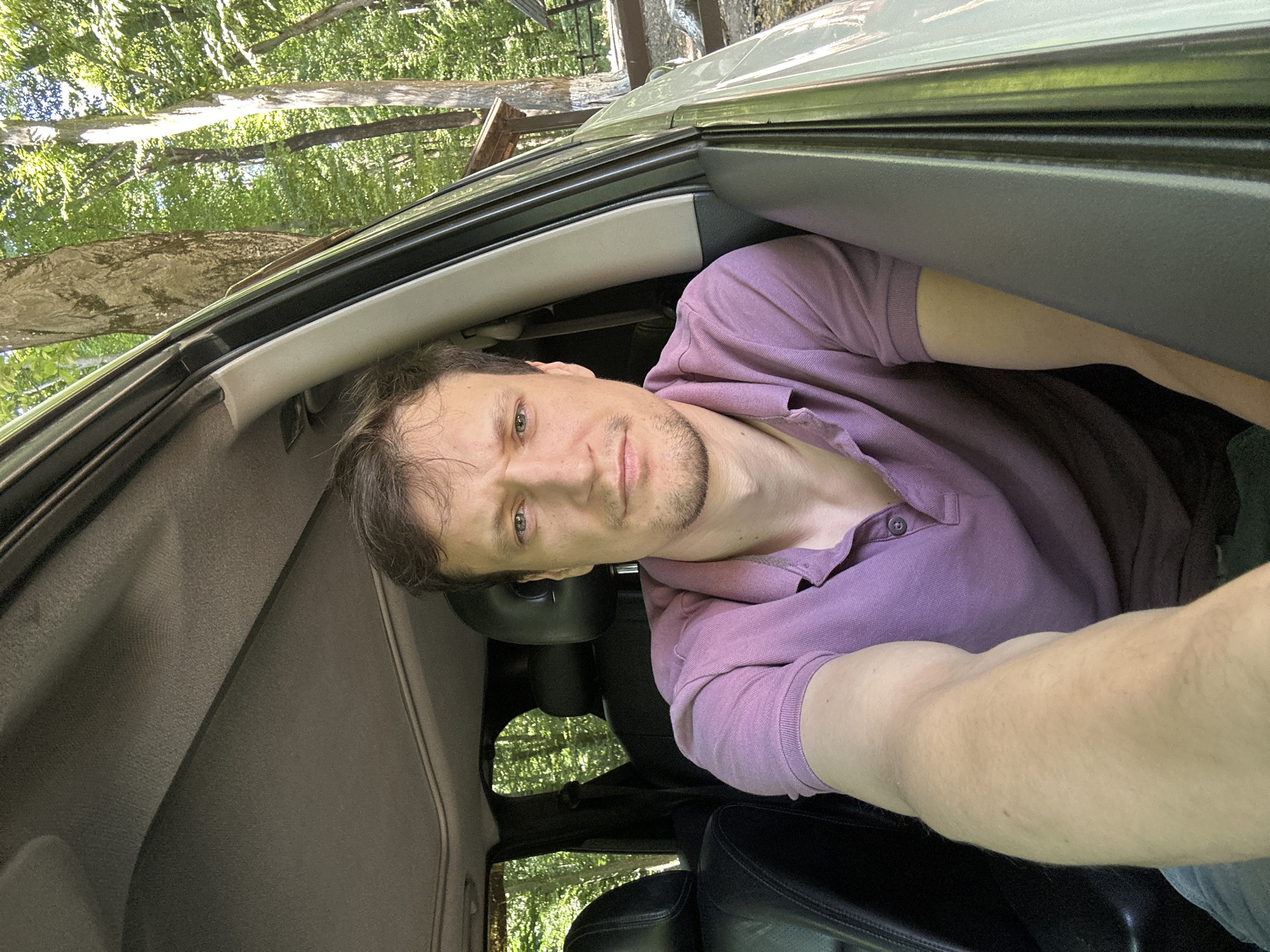 A man is taking a selfie in the driver's seat of a car, parked outdoors with trees and foliage visible through the open car door and windows. He has brown hair and is wearing a purple polo shirt. The interior of the car shows black leather seats and a grey headliner. The environment appears to be a wooded area, possibly a park or roadside. The location is the village of Shatava, Ukraine.