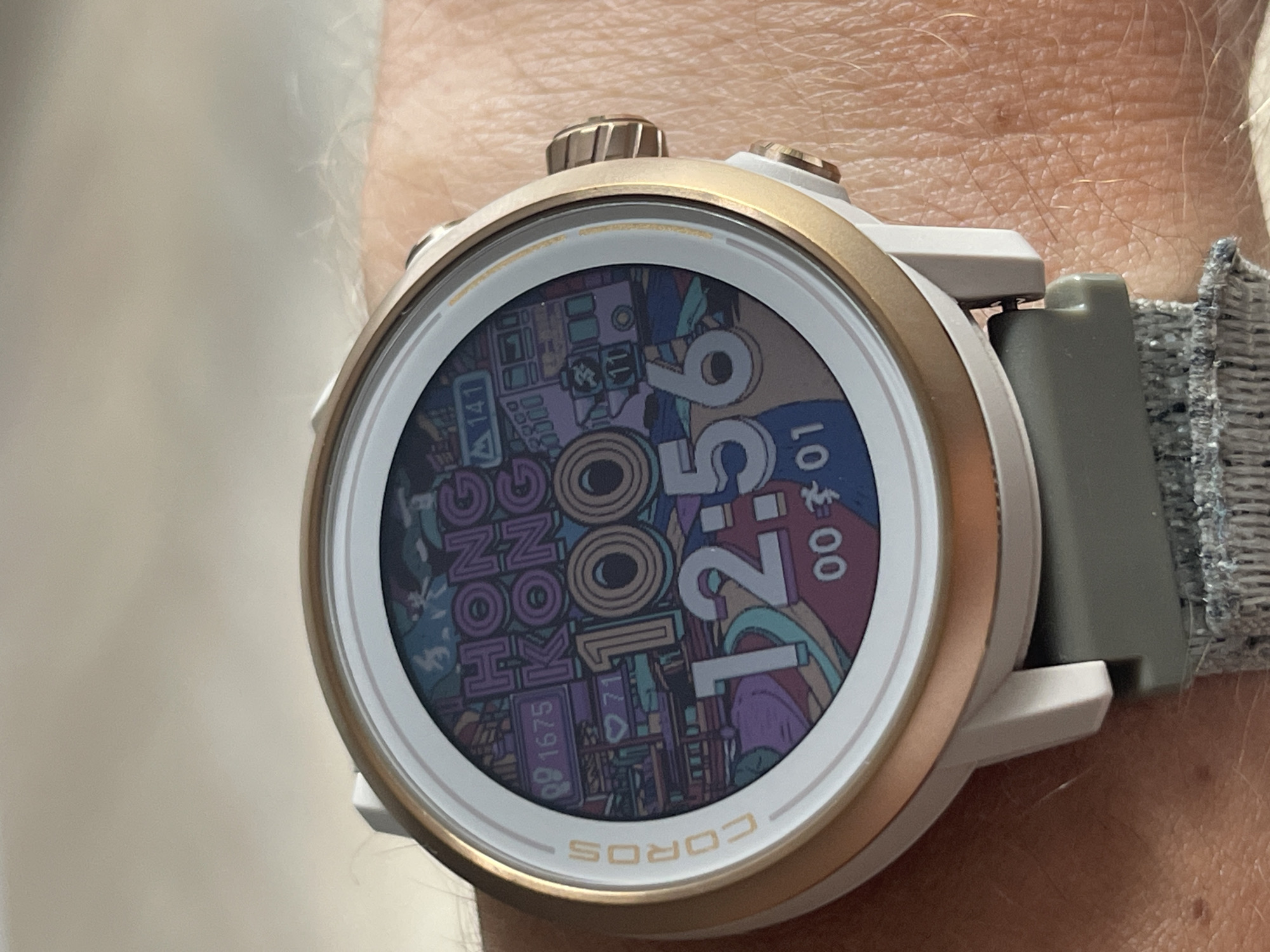 A close-up, high-angle shot shows a COROS brand smartwatch on a person's wrist. The watch face displays a colorful, cartoon-style graphic with the words "HONG KONG" in large purple letters and "100" in orange. Below this, the time is displayed as "12:56". Small icons for steps, heart rate, and other metrics are scattered around the face. The watch has a white casing with a gold bezel and a grey strap. The background is blurred, showing a lightly textured, light-colored surface, likely skin.