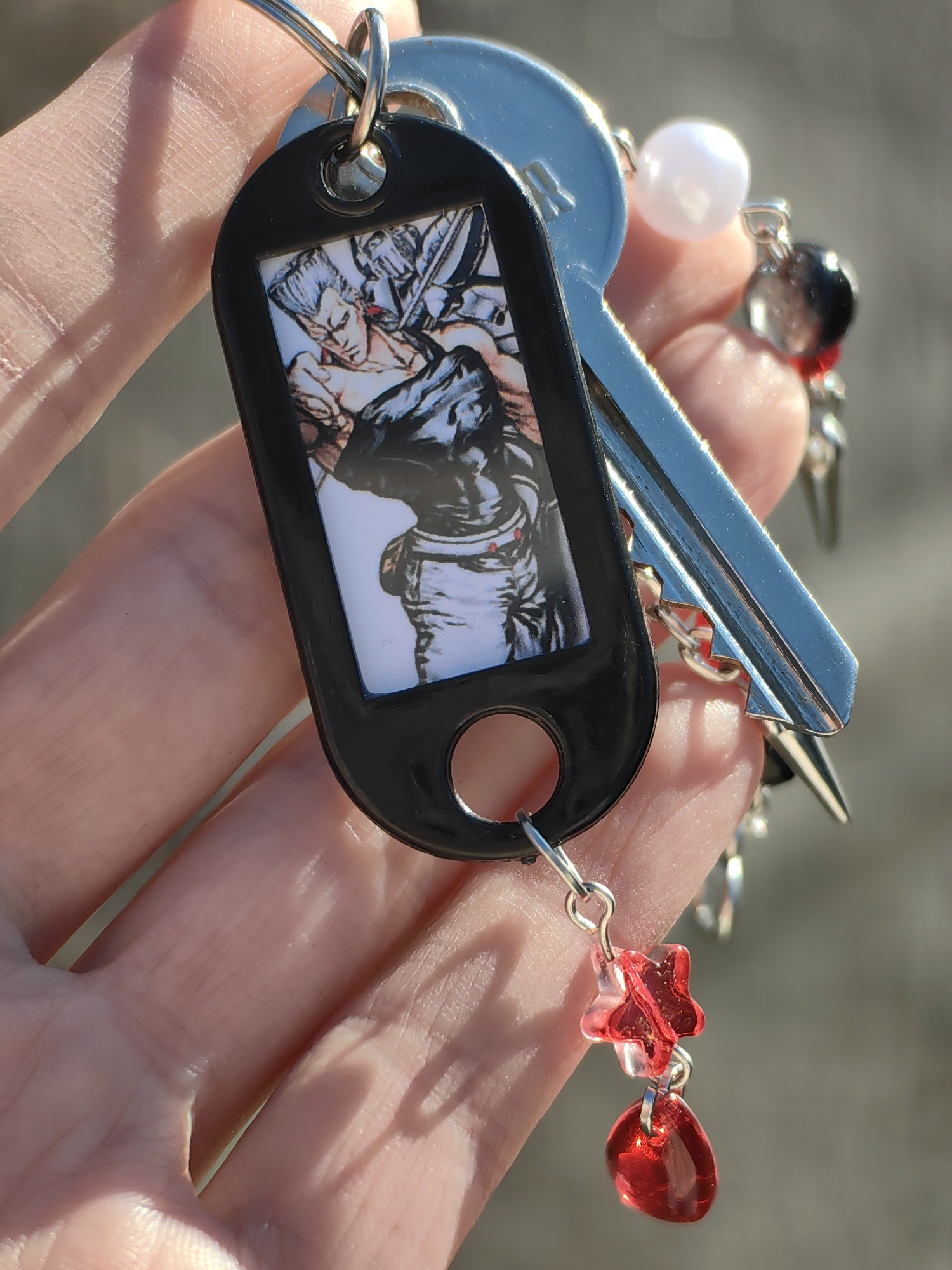 An adult human hand holds a set of keys and keychains in Chaikovsky, Russia, under bright sunlight. The hand, visible with detailed skin texture and fingerprints, supports a metallic split key ring. Attached to this ring is a prominent black, oblong key fob featuring a rectangular inset image. The image is a colored illustration depicting two male figures in a dynamic, possibly confrontational pose, rendered in an anime-like style; one figure on the left is bare-chested, while the other on the right wears dark clothing. Below the image, the fob has a circular cutout from which a small chain extends, connecting to two translucent red charms: a star-shaped charm and an oval or tear-drop shaped charm. Also on the key ring is a single, shiny silver-colored key with a complex bitting pattern, reflecting the sunlight. A light pink, spherical bead charm is also attached. In the blurred background, additional metallic items, including what appears to be another key or spiked charm, and more small red beads are partially visible. Strong shadows are cast from the fingers onto the palm, indicating the direction of illumination from an overhead light source. The background is a soft, out-of-focus mix of grayish and light brown tones, suggesting an outdoor environment.
