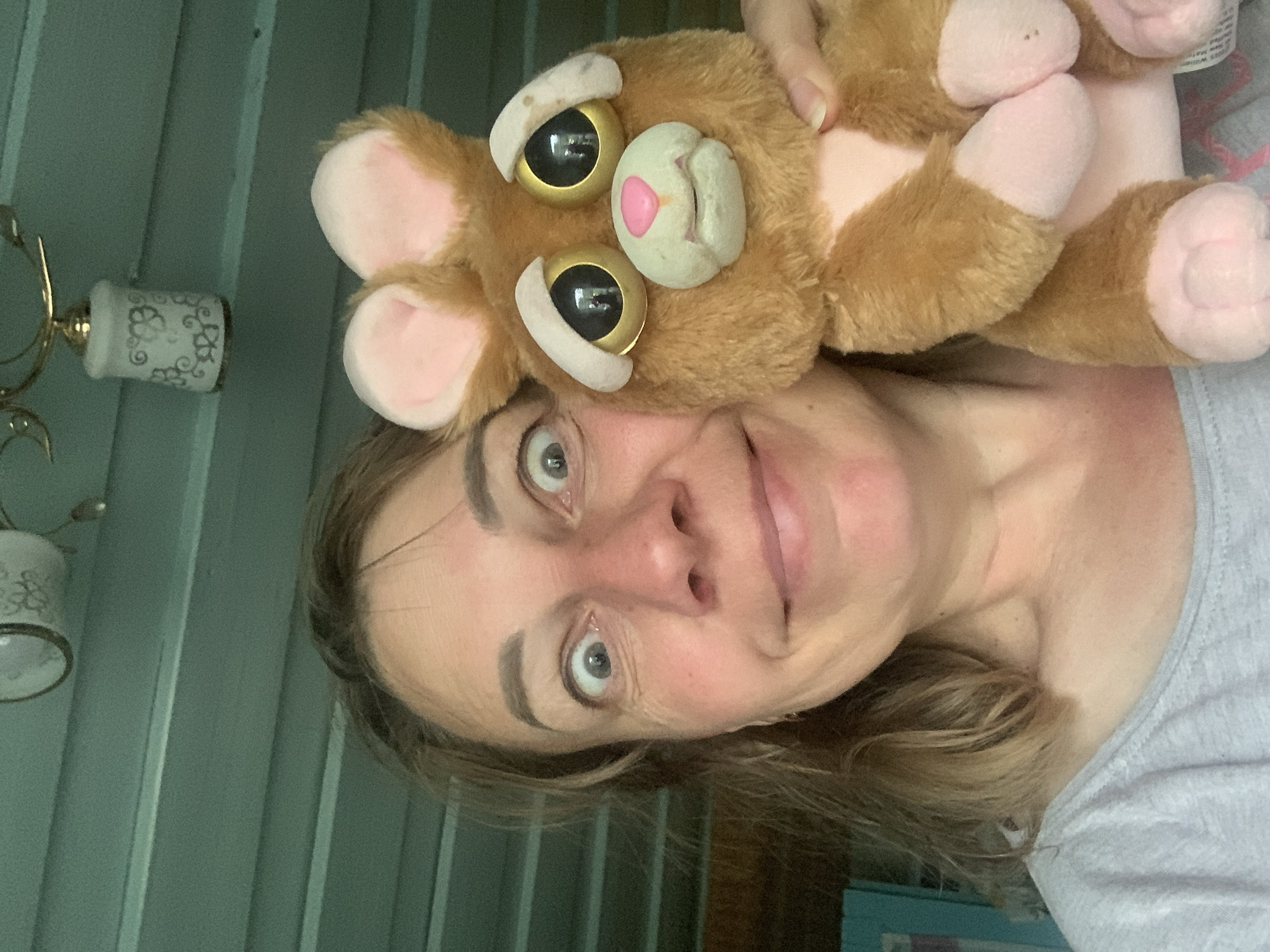 A woman is holding a stuffed toy animal in front of her face, with both of them looking at the camera. The woman has wide, surprised eyes and a slight smile. The stuffed animal is brown with large, round eyes and pink ears. They appear to be indoors, with a green, slatted wall behind them and a light fixture visible in the upper portion of the frame. There is no discernible indication of the location being in Відзаўскі сельскі Савет, Belarus.