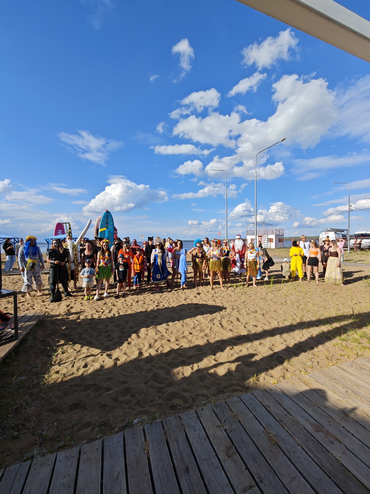 On a sandy beach in Chaikovsky, Russia, a large group of people, some in costumes, stand facing the camera under a bright blue sky with scattered clouds. The individuals are gathered near the water, with surfboards and other beach equipment visible behind them. A wooden walkway extends into the foreground, casting shadows on the sand. In the background, there are streetlights, a lifeguard tower, and a few vehicles. The overall atmosphere appears festive and relaxed.