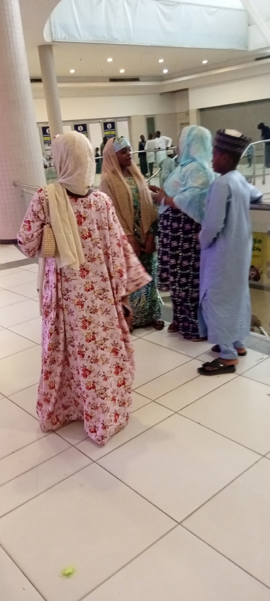 In Gandu Albasa, Nigeria, a group of people, including men and women in traditional Islamic attire, are gathered indoors, possibly within a mall or community center. The women are wearing hijabs and flowing dresses, with one woman in a pink floral-patterned abaya and a beige scarf draped over her shoulders. Another woman is in a light blue hijab and a patterned skirt. A young boy stands to the right, dressed in a light blue kaftan and wearing a patterned cap. The environment features tiled flooring and overhead lighting. The individuals appear to be conversing or standing together.