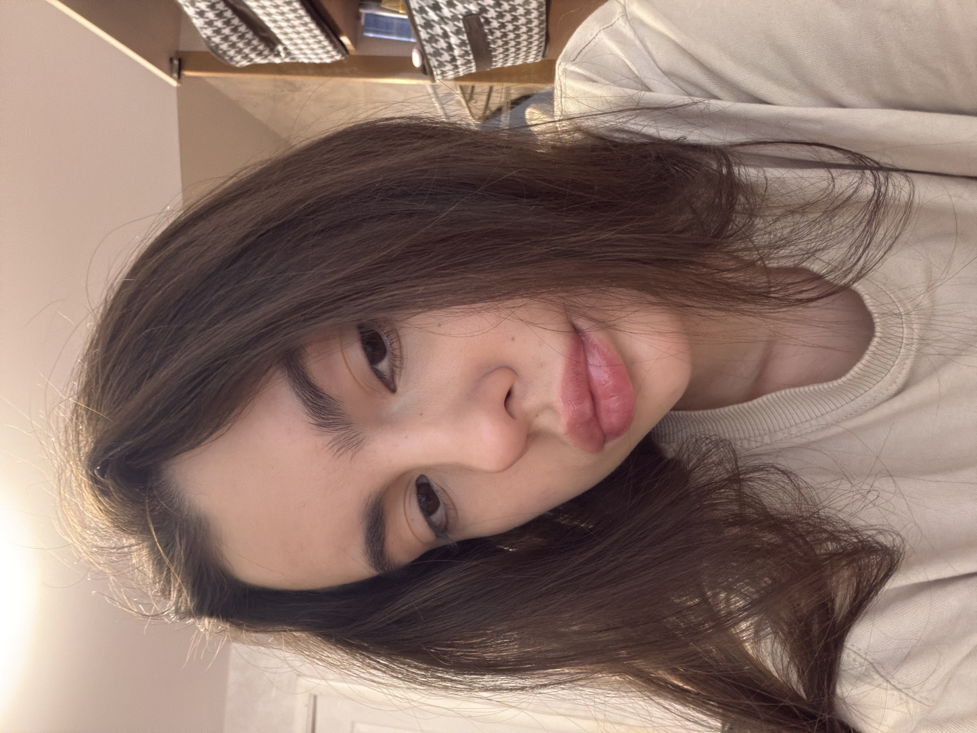 A close-up, rotated selfie of a young person with dark brown hair and fair skin. They are wearing a light beige crewneck sweater. The background is blurry, showing a shelf with items and a houndstooth pattern. The setting appears to be indoors. The location is not discernable as Chaikovsky, Russia from the provided visual information.