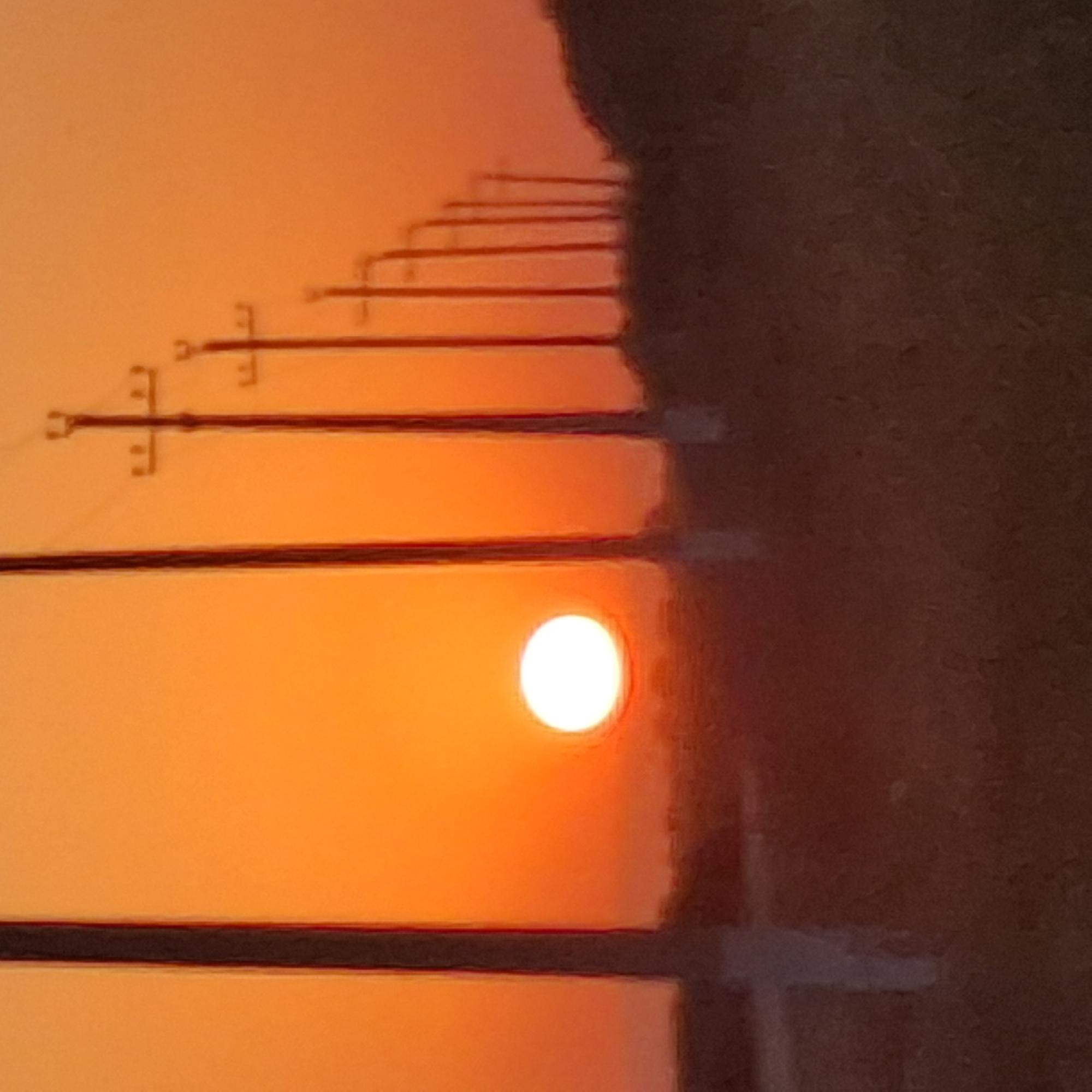 A wide shot captures a vibrant sunset in Қарааспан, Kazakhstan. The sun, a brilliant white orb, is positioned low on the horizon, casting an intense orange glow across the sky. A row of utility poles, silhouetted against the sunset, march across the frame from left to right. The poles are dark and slender, with crossbars visible on some, suggesting they carry electrical wires. In the foreground, the ground is covered in dry grass and low vegetation, rendered in muted browns and reds by the sunset light. The overall atmosphere is one of serene twilight.
