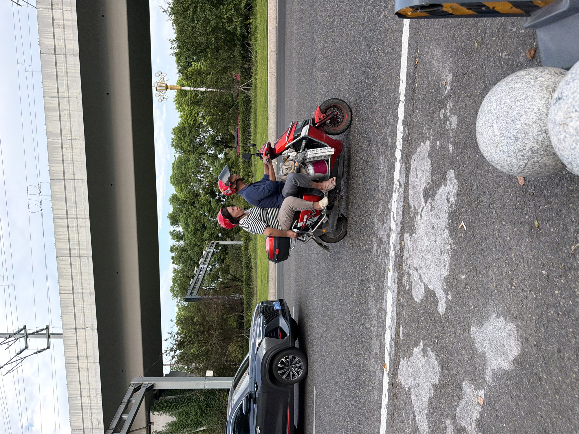 The image depicts a street scene in Jiangjin, China, under an elevated highway structure. A dark gray SUV is parked on the left side of the road. On the right, a man and a woman are riding a red electric scooter. Both are wearing red helmets. The woman is seated behind the man, wearing a striped shirt and light-colored pants. The man is wearing a dark blue shirt and gray pants. The scooter has a black storage box on the rear and several items attached to the front, including buckets and what appear to be tools. The road surface is asphalt with white lane markings. In the background, there are trees and a street lamp with multiple lights. The sky is overcast.