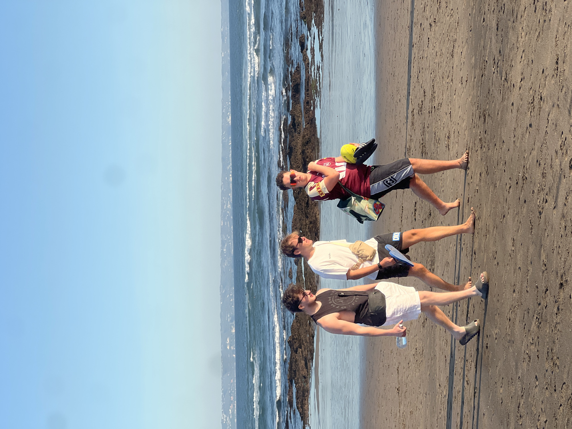 Three young men are walking on a sandy beach near the ocean. The man in the front is wearing a black tank top with "GYM" visible on it and white shorts. He is carrying a clear water bottle in his left hand. He has on dark grey sandals. The middle man is wearing a white t-shirt and black shorts. He has a light beige fanny pack around his waist and is holding a light blue paddle in his right hand. The man in the back is wearing a maroon and white jersey with "SHER" visible on the back and black shorts. He has on sunglasses and is carrying a yellow ball and a pair of black sandals in his arms. The ocean has waves crashing against rocks along the shore. The sky is clear and blue. The sun is shining brightly, suggesting it is daytime. The scene appears to be a casual outing or a trip to the beach.
