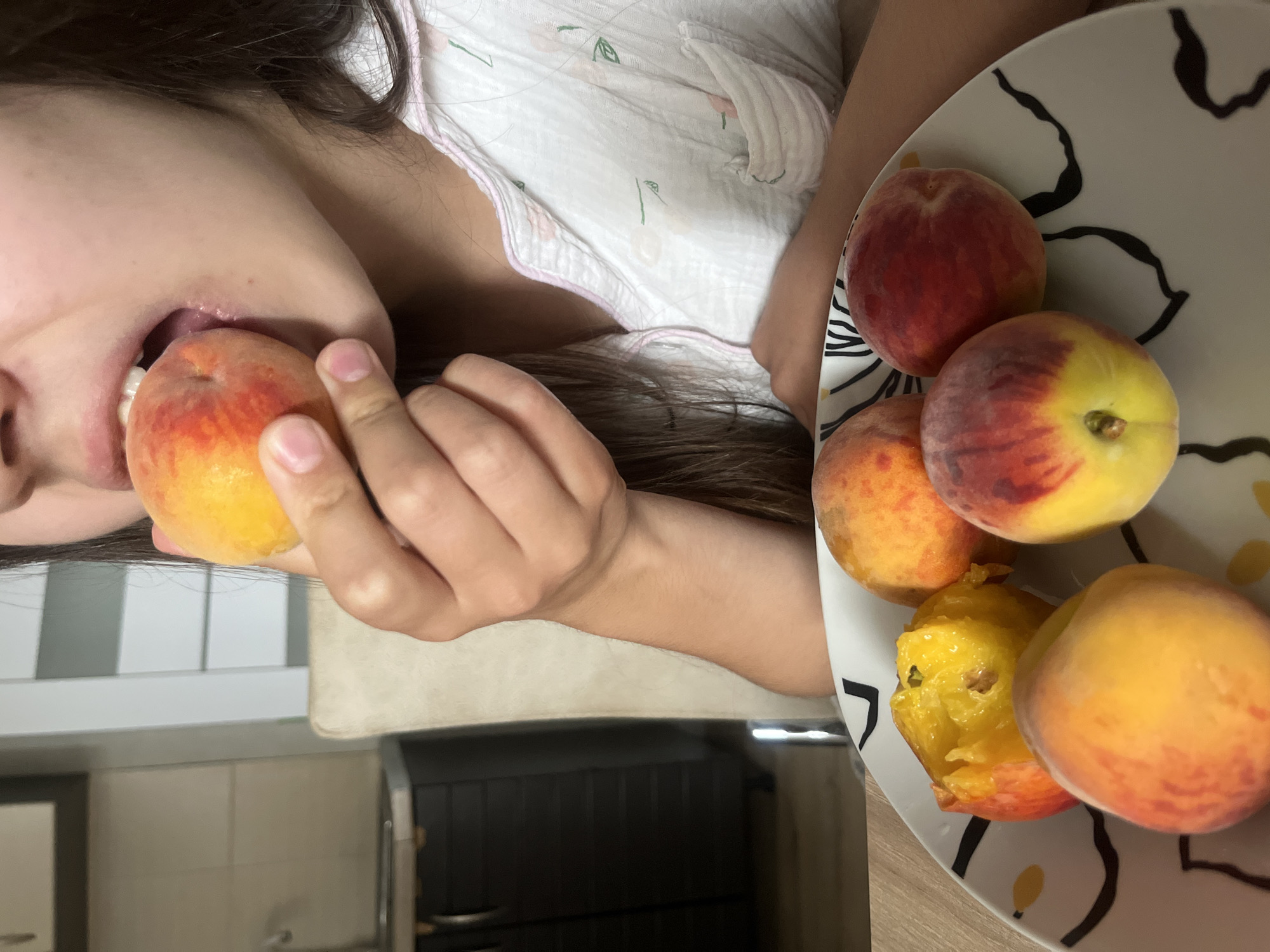 A person is eating a peach, holding it in their right hand and bringing it to their mouth. The person is wearing light-colored pajamas with a floral pattern. In the foreground, there is a plate with several peaches. One peach on the plate has been partially eaten. The background shows a kitchen area with cabinets and a countertop. The overall scene suggests a casual, domestic setting, possibly a home in Rybnitsa, Moldova.