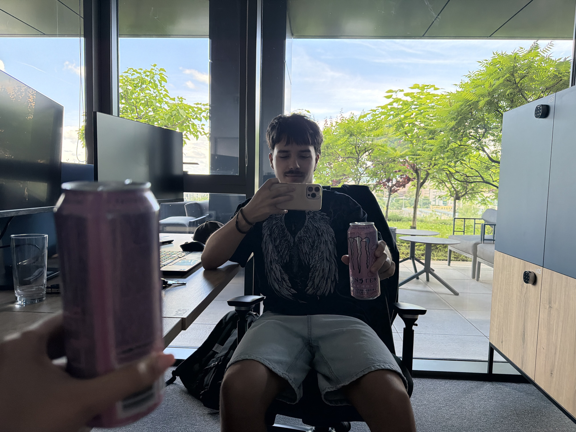 The image portrays a casual scene within a modern office or workspace, likely located in Sofia, Bulgaria. A young man, featuring short dark hair and a mustache, is seated in a black office chair, facing forward. He is dressed in a black t-shirt with a winged graphic design and light-colored shorts.
In his right hand, the man holds a light-colored smartphone, appearing to take a selfie or record. His left hand grasps a pink Monster Energy drink can, clearly labeled "MONSTER ENERGY," "SUGAR FREE," and "STRAWBERRY." Another identical pink Monster Energy can is held in a blurred hand in the immediate foreground on the right side of the image, suggesting a shared experience or a focus on the beverages.
The man is positioned at a wooden desk where two dark computer monitors are visible, along with a glass, a laptop, and a set of keys. A black backpack is placed on the floor beside his chair. To his right, a contemporary storage unit made of wood and dark grey panels features a black digital lock marked "G26."
Beyond the large windows, bright daylight illuminates a vibrant outdoor scene. Lush green trees are prominent against a blue sky dotted with white clouds, indicative of a pleasant late morning or afternoon. Outdoor patio furniture, including light grey tables and chairs, can be seen, contributing to the open and modern aesthetic of the setting.