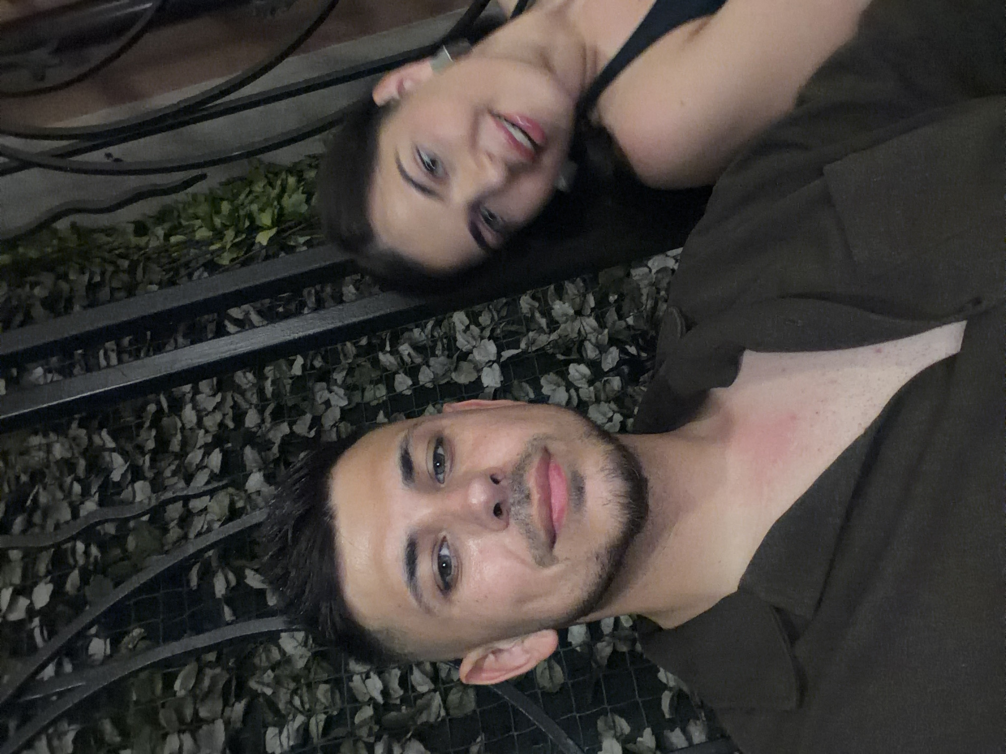 In the city of Sozopol, Bulgaria, a close-up, low-angle, indoor selfie shows two individuals. A man with short, styled dark hair and stubble is in the foreground, facing the camera with a slight smile. He is wearing a dark brown button-up shirt with the collar open, revealing the top of his chest. To his right and slightly behind him, a woman with long dark hair is partially visible, smiling and looking away from the camera. She is wearing a dark tank top and large, square earrings. The background features a dark metal fence or grate with artificial green foliage behind it, suggesting an outdoor or semi-outdoor setting, possibly a restaurant or bar area in Sozopol.