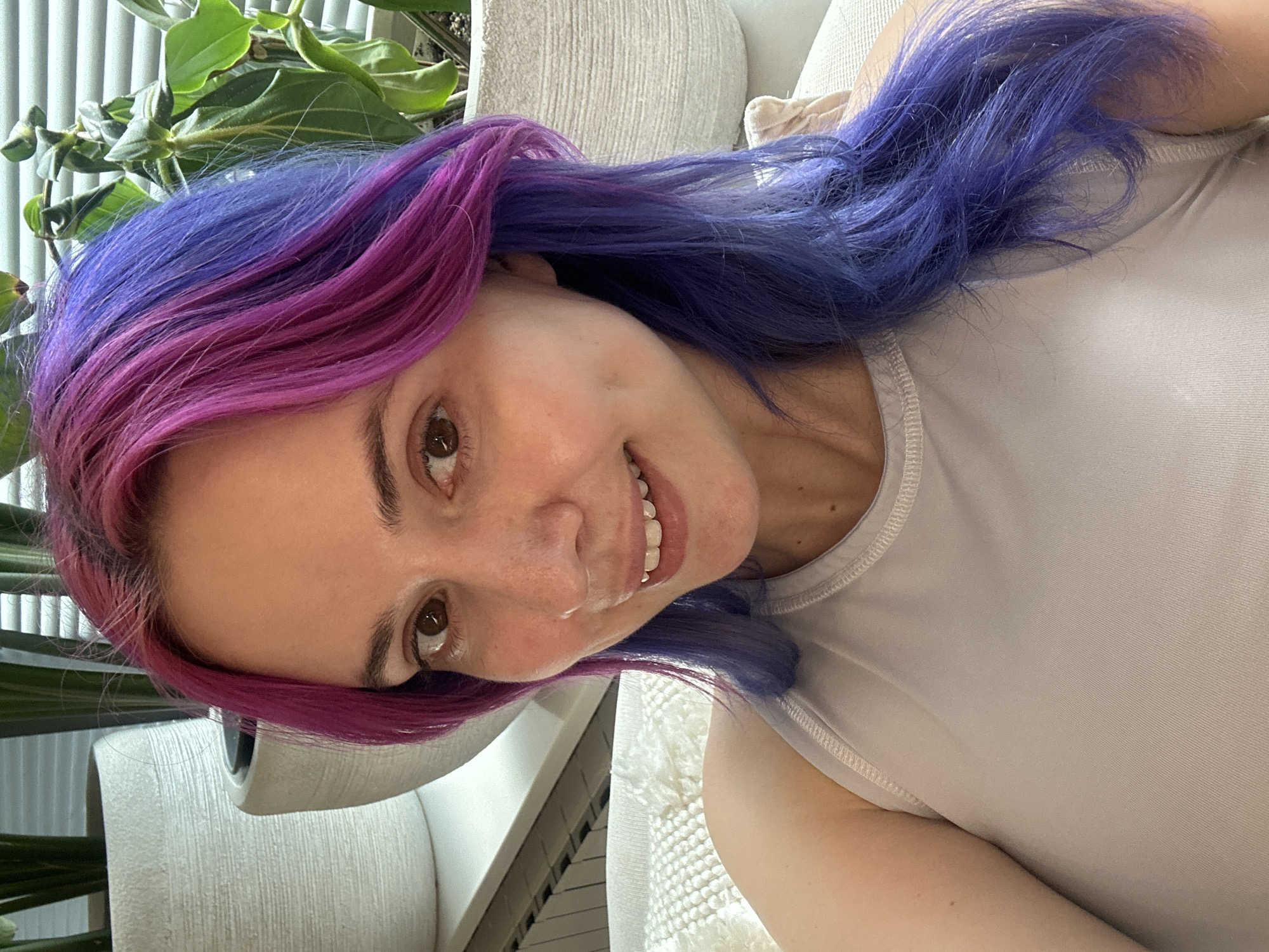 A casual portrait captures a smiling woman with distinctive two-tone hair, dyed vibrant magenta on one side and deep blue on the other. Her head is tilted, and she looks directly at the viewer with warm, brown eyes. Her smile is broad, revealing white, even teeth, and her expression conveys friendliness and contentment. Freckles are visible across her nose and cheeks.
She is wearing a light-colored, possibly off-white or light grey, sleeveless top with a ribbed texture around the neckline. The setting appears to be indoors during daytime, suggested by the natural light reflecting in her eyes and illuminating the scene. In the background, green foliage from an indoor plant is visible, along with textured light-colored furniture or a wall, indicating a comfortable home or office environment. There are no specific visual cues within the image to confirm the provided location context of Zagreb, Croatia. No text is visible in the image.