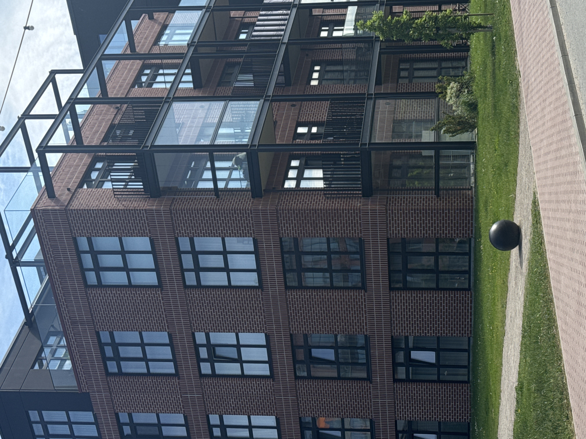 A modern brick building with black framed windows and balconies is shown in this image taken from a street level perspective. The building has several floors and features a contemporary architectural style with extensive use of glass and metal accents. A dark, spherical sculpture sits on the grass in front of the building. The scene appears to be in an urban setting, with a paved walkway and a street visible in the foreground, and lush green grass surrounding the sculpture. The sky above is partly cloudy. The building's design and materials suggest a relatively new construction. The image has been identified as being in Tallinn, Estonia.