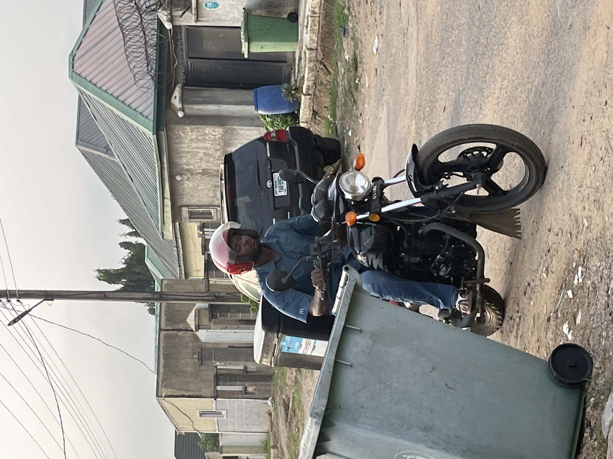 A man wearing a pink helmet, blue denim shirt, and blue jeans is riding a black motorcycle. He is positioned in front of a large, green, commercial trash container. Behind the motorcycle is a dark SUV with license plate starting "LA". Further in the background, there are residential buildings with various exterior designs. Overhead power lines are also visible. The location appears to be in an urban environment, likely in Abuja, Nigeria. The setting suggests a street or roadside scene.