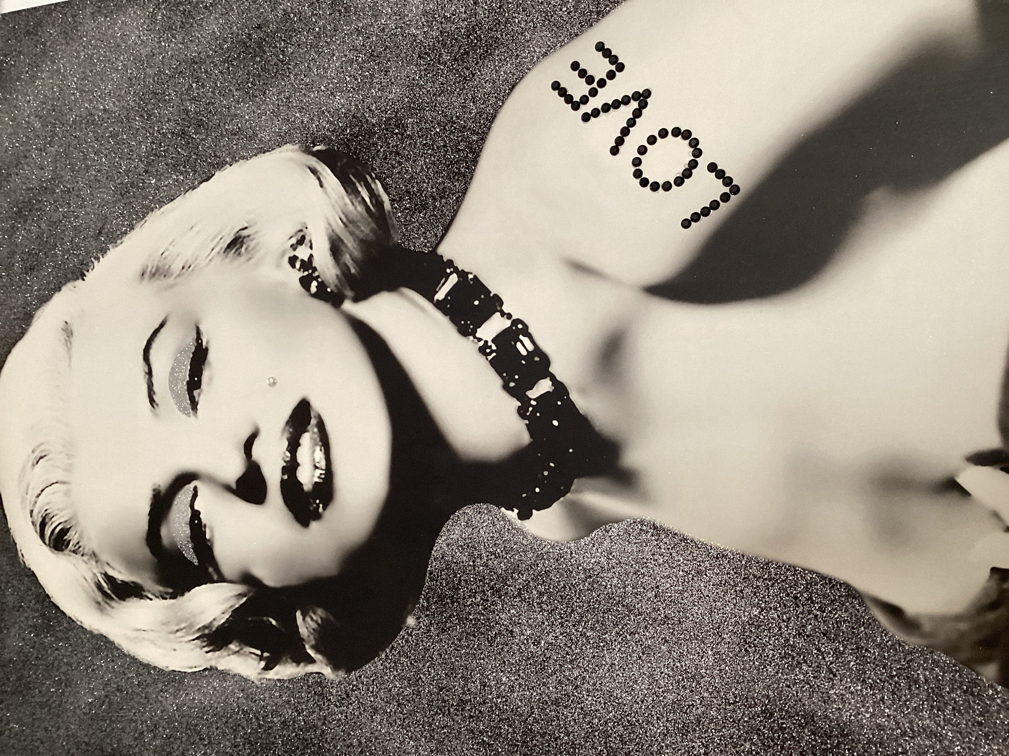 The image is a black and white portrait of Marilyn Monroe. She is looking directly at the camera with a sultry expression. Her eyes are emphasized with glitter, and she has a mole on her cheek. She is wearing a dark, beaded necklace. On her right shoulder, the word "LOVE" is written in small, dark dots. The background is speckled and appears to be a textured surface, possibly glitter or sequins. The image is likely a piece of art or a poster. There is no information about the city of Blanes, Spain, in this image.