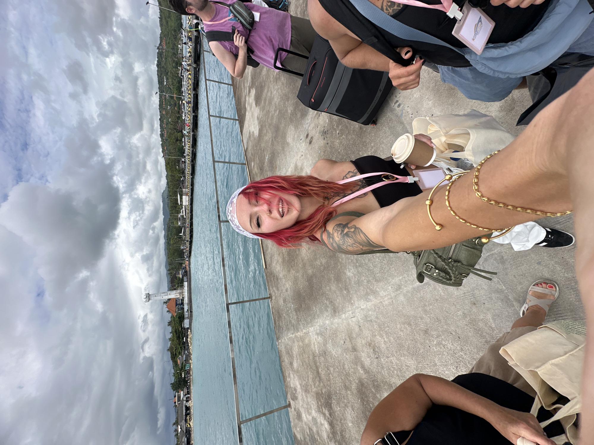 A group of people are on a concrete pier next to a body of water. A fair-skinned person with pink hair, a white lace headband, and tattoos on their arm is holding up a coffee cup and smiling. This person is wearing a black top and a pink lanyard. In the foreground, another person's arm with gold bracelets is visible, as well as a person's leg wearing a tan sandal. The person is also sitting next to an olive-colored purse. Two other people are standing with luggage. In the background, a view of Kampung Toyapakeh, Indonesia is seen, with buildings, a lighthouse, and cloudy skies. The concrete pier has a metal railing.