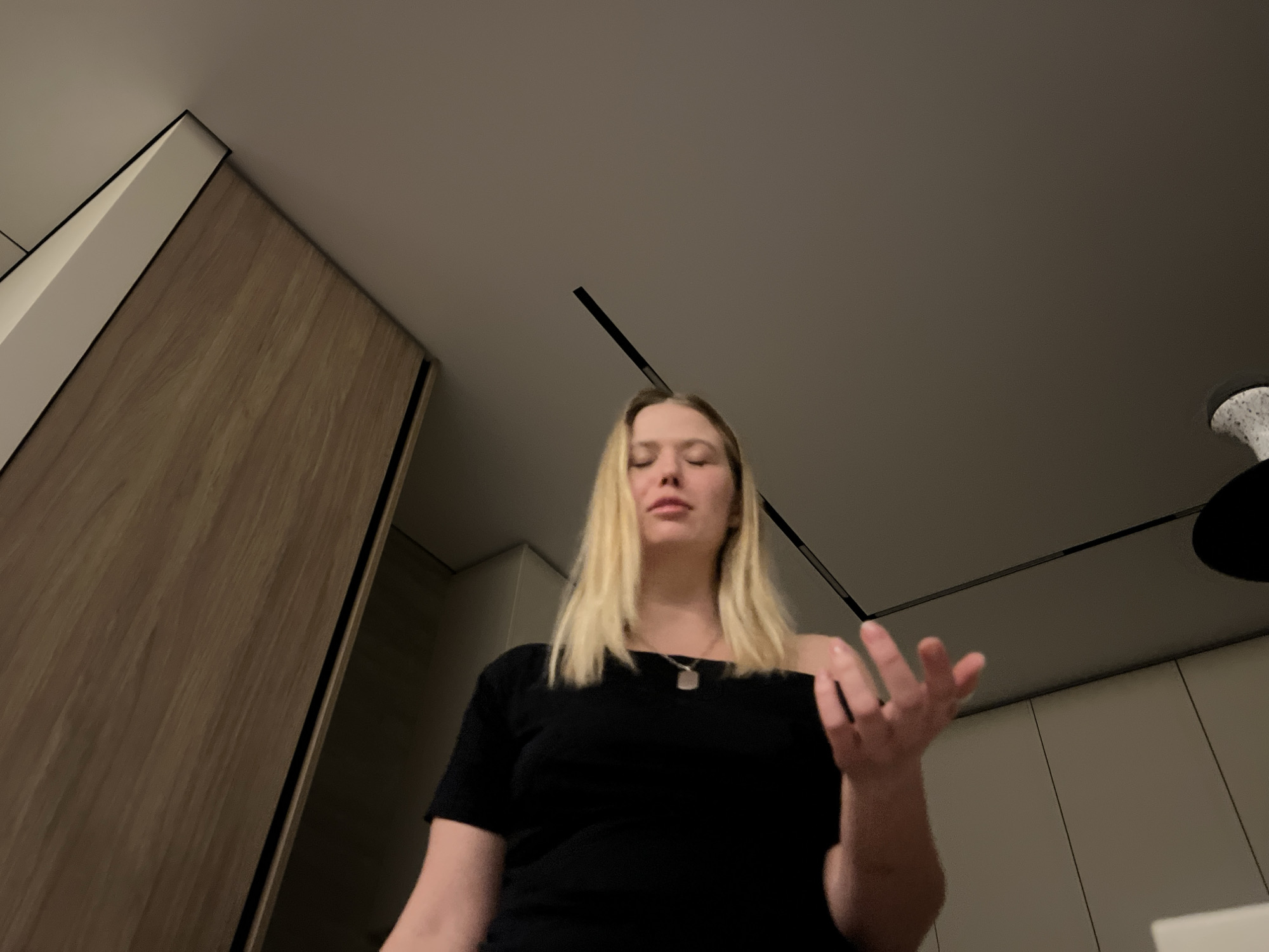 A woman with blonde hair, wearing a black shirt and a necklace, is shown from a low angle. She appears to be indoors, possibly in a room with a ceiling fan and wooden cabinets. The precise location is not ascertainable within Рубцово, Russia.