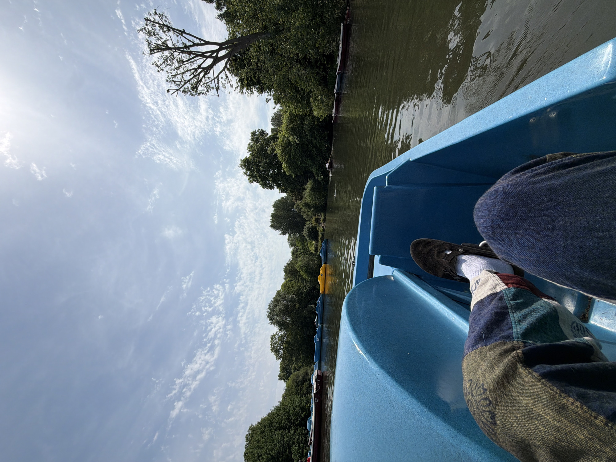 A low-angle, partially obscured, point-of-view shot shows a person in a blue pedal boat, likely on a body of water. The right side of the image features the side and front of the boat, constructed of light blue molded plastic. A leg, clad in patchwork denim pants, a white sock, and a dark shoe, rests in the boat. The background shows a narrow body of water bordered by trees on either side, with several moored boats visible along the shoreline. Above, a bright sky with scattered clouds and a hint of the sun is visible. Given the presence of pedal boats and the landscape, the location is likely a park in London, United Kingdom, possibly Hyde Park or Regent's Park. The image conveys a sense of leisure and recreation.