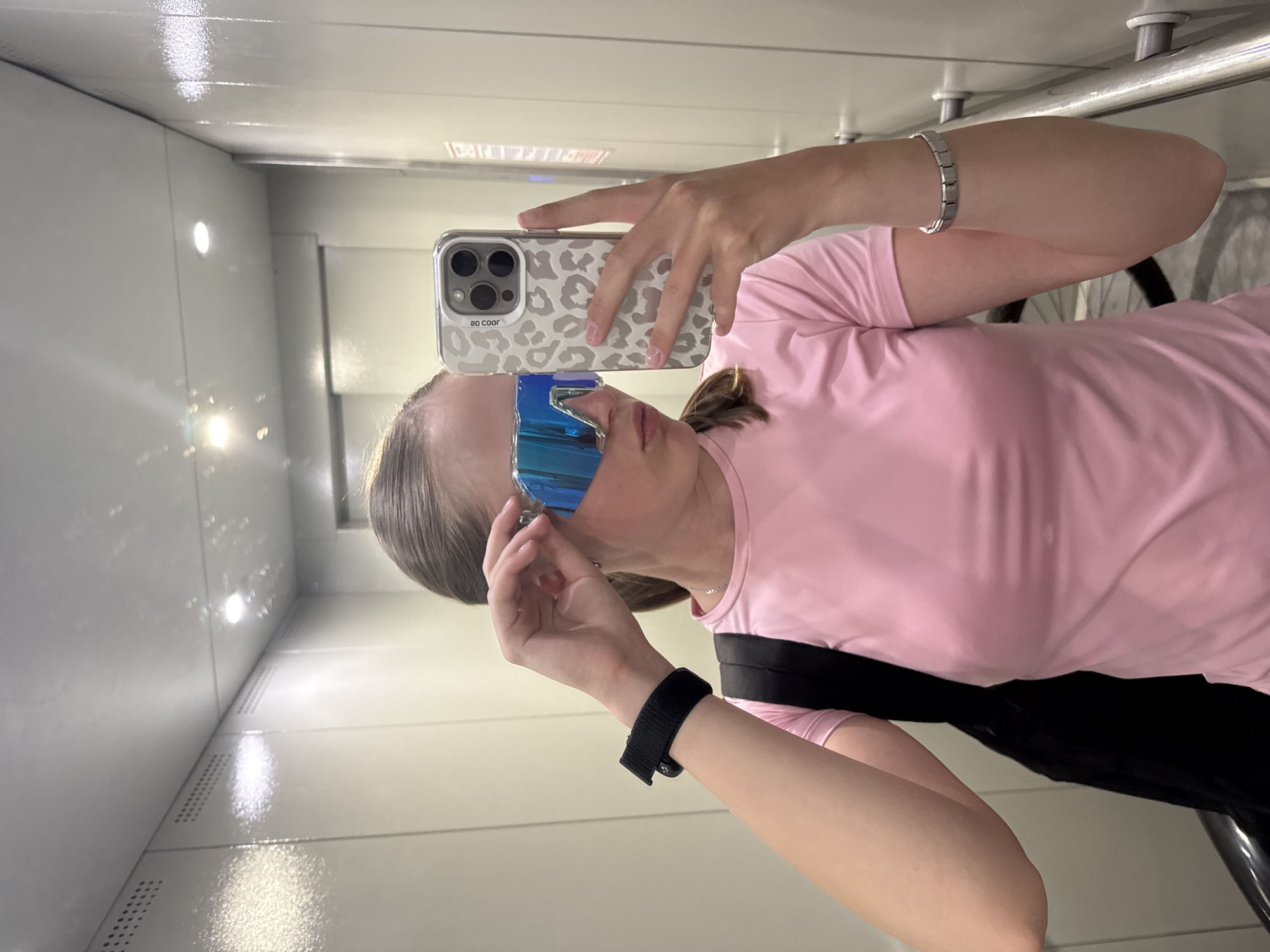 A young person is taking a selfie in what appears to be an elevator or a small, enclosed space with white walls and a reflective surface, possibly a mirror or polished metal. The person is wearing a pink shirt and black undergarment strap. They have on a silver bracelet on their left wrist and a black sports watch on their right wrist. The person is holding a smartphone with a light-colored, patterned case to take the selfie. The person is also wearing blue, futuristic-looking sunglasses. The selfie is reflected in the shiny surface around them, creating an impression of depth. The background is generic, suggesting a modern, perhaps utilitarian, interior space. There is no explicit evidence linking this image to Chaikovsky, Russia.