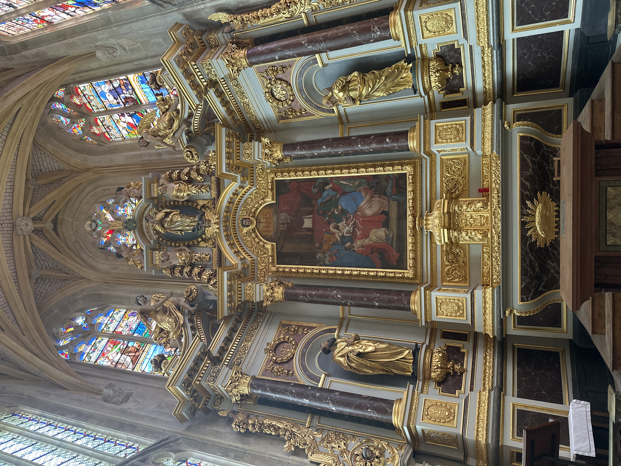 The image shows the interior of a church in Dieppe, France, specifically the altar area. A large, ornate altarpiece dominates the scene, featuring a central painting depicting a religious scene with several figures. The altarpiece is adorned with intricate gold detailing, marble-effect columns, and sculptures of saints or angels. Above the altarpiece, stained-glass windows with colorful religious imagery are visible, casting light into the space. In the foreground, an altar table is present. The architectural style of the church, with its high ceilings and vaulted arches, is also evident.