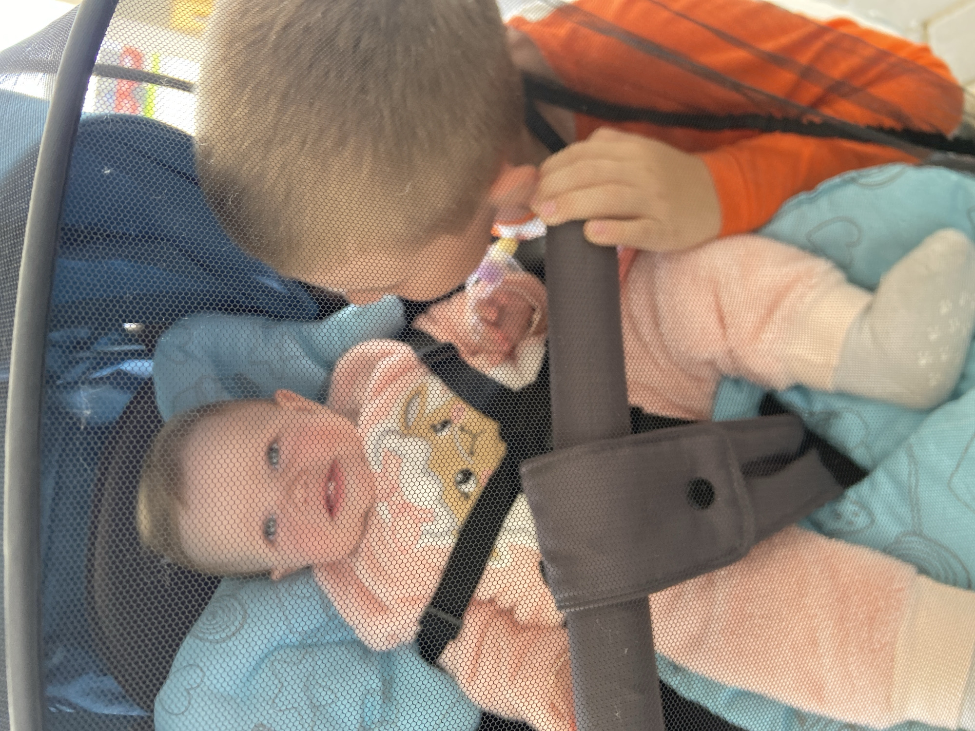 The image depicts two children viewed through a fine, dark mesh netting, likely the cover of a stroller or playpen. An infant, with light hair and fair skin, is seated in what appears to be a light blue padded stroller insert or blanket featuring faint, lighter-toned patterns. The infant wears a light pink long-sleeved outfit with a central light-colored cartoon animal graphic, possibly a bunny, on the chest. The child is secured by dark gray safety straps and a padded buckle system, and is wearing light-colored socks or booties. The infant's mouth is open in what appears to be a smile, revealing lower teeth, and the gaze is directed forward.
To the right, partially visible, is a second child, a toddler or young child with light brown hair and fair skin, wearing a long-sleeved orange shirt that may have subtle darker vertical stripes. This child is leaning towards the infant, with their right hand grasping a horizontal dark gray bar of the stroller frame. Their left hand holds a small, colorful object, possibly a pacifier or toy, near the infant's face. The background, viewed through the netting and behind the children, is largely out of focus but suggests an indoor or sheltered setting with varied light and dark elements. The image does not contain any discernible features or metadata to indicate the location in the city of Chaikovsky, Russia, or any other specific geographical location.