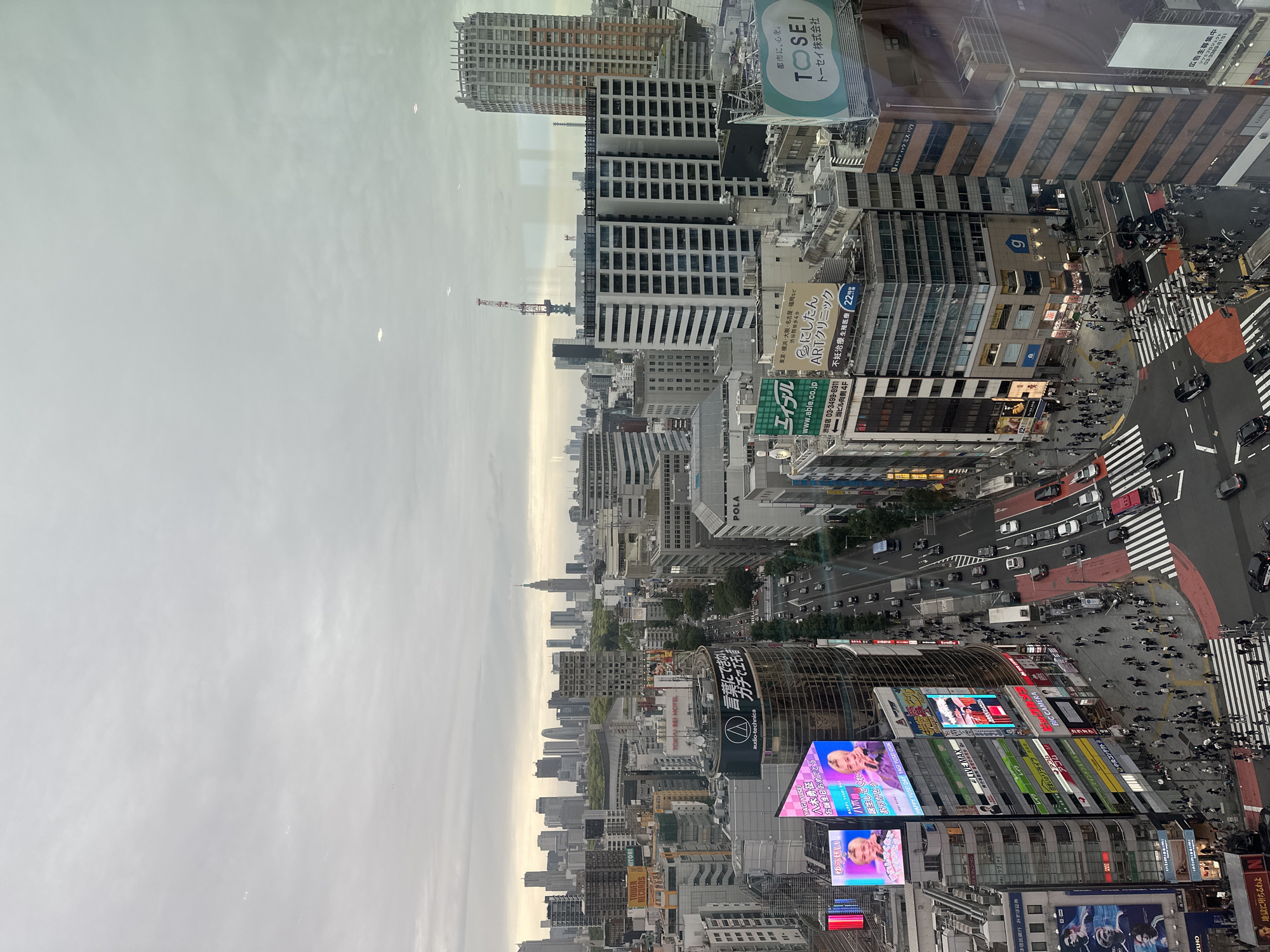 The image shows an elevated view of the Shibuya district in Tokyo, Japan, on a cloudy day. The famed Shibuya Crossing is visible, filled with pedestrians. The scene includes numerous high-rise buildings covered with billboards. Prominent signs include those for BIC CAMERA, audio-technica, and ToSEI. The Tokyo skyline is visible in the background.