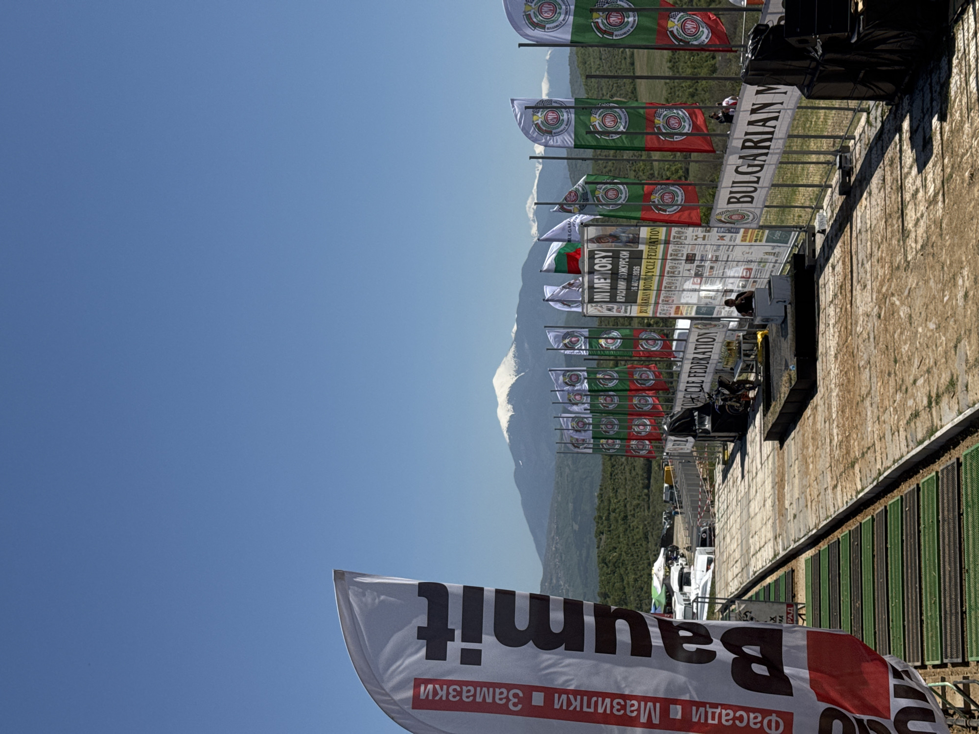 The image captures a scene at what appears to be an outdoor event in the city of Selishte, Bulgaria, judging from the presence of Bulgarian flags and signage in Bulgarian. The foreground is dominated by a "Baumit" promotional flag, suggesting a construction or building materials company is a sponsor. A series of Bulgarian Motorcycle Federation flags are aligned in the background, with banners dedicated to the memory of Krasimir Bozhurski. A mountainous landscape is visible in the distance, partially obscured by the event infrastructure. People are present but not distinctly identifiable. The image appears to be taken from a slightly elevated perspective, looking towards a raised platform or structure. The sky is clear and blue, indicating fair weather. The environment suggests an organized public gathering.