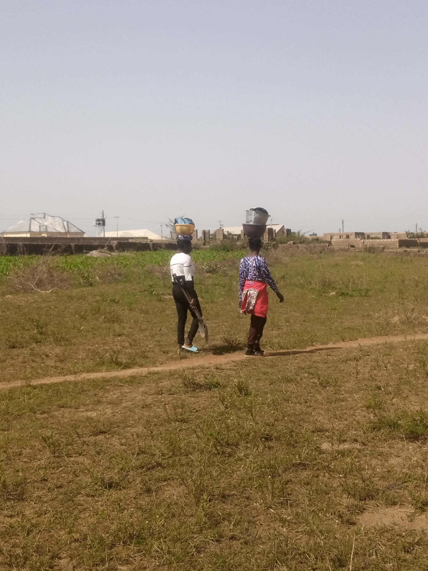 The image depicts an outdoor scene in Kwang, Nigeria, featuring two individuals walking across a dry, grassy field under a clear sky. The ground is uneven and covered with sparse, brown vegetation, indicating arid conditions. A faint dirt path runs diagonally across the foreground, suggesting a well-traveled route.
In the middle ground, a person on the left is walking away from the camera, carrying a yellow basin filled with items on their head. They are dressed in a white long-sleeved shirt and black trousers. They also hold a shovel in their right hand. To the right, another person is also walking away from the camera, carrying two buckets on their head, one atop the other. This individual wears a blue and white patterned long-sleeved shirt and a red skirt or wrap around their waist. They are also dressed in dark trousers and dark shoes.
In the background, there are several structures. To the left, a building with a metallic roof is visible, possibly under construction or renovation. Further back, a series of brick structures, some appearing incomplete, are scattered across the landscape. A utility pole with wires can be seen among these structures, and a hazy, light-colored horizon suggests a dusty or distant urban or semi-urban environment. The overall atmosphere is one of a rural or developing area, likely experiencing warm weather given the dry vegetation and clear sky.