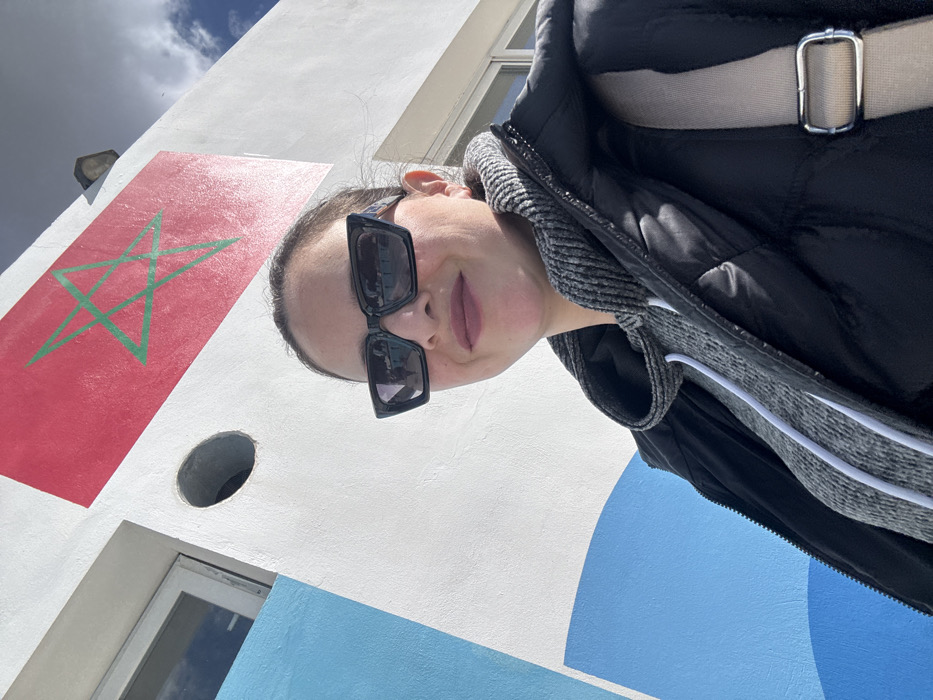 A female individual is captured in a close-up, upward-angled photograph, consistent with a selfie, positioned against the exterior wall of a building in Meknes, Morocco. She faces slightly towards the camera with a faint smile, wearing dark, rectangular sunglasses that reflect the sky, dark lipstick, a grey textured hoodie, and a black quilted puffer jacket. A beige shoulder strap from a bag is visible over her right shoulder. The white-painted building wall behind her features a large, painted Moroccan flag, characterized by a red field with a green pentagram, in the upper right section. To the left of the flag, a dark circular opening is present. Further to the left and slightly higher, a rectangular window with a light-colored frame reflects the sky. A curved blue design is painted on the lower left portion of the white wall. Patches of blue sky with white clouds are visible at the top right of the frame.