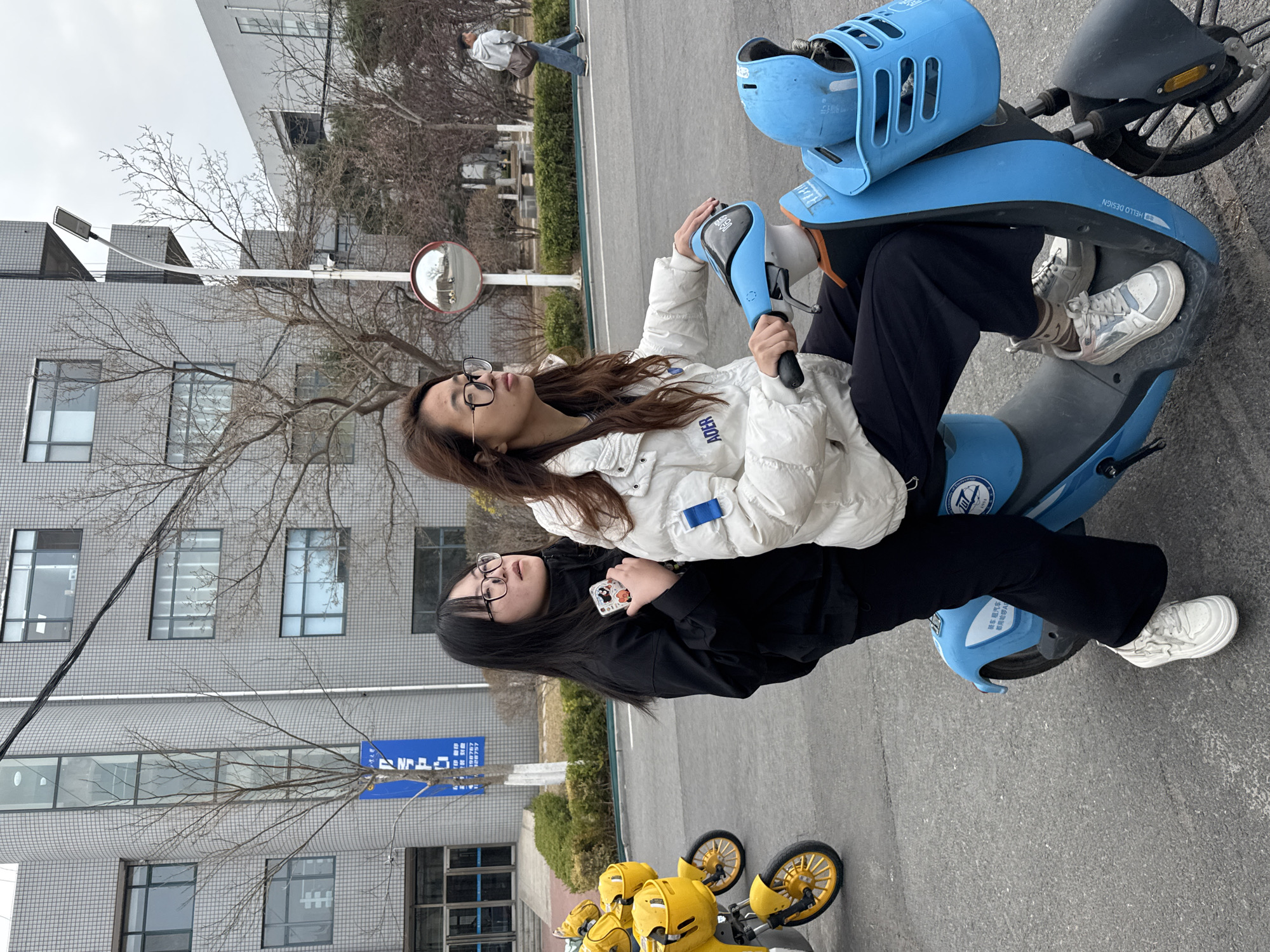 Two young women, one wearing a white puffer jacket and the other a black jacket, are sitting on a light blue electric scooter in an urban environment. The woman in the white jacket is operating the scooter. The scooter has a helmet holder on the front. The backdrop includes a multi-story building with windows, a few bare trees, and other parked yellow bikes. A sign with Chinese characters is also visible. The location is the city of Zhongtun, China.