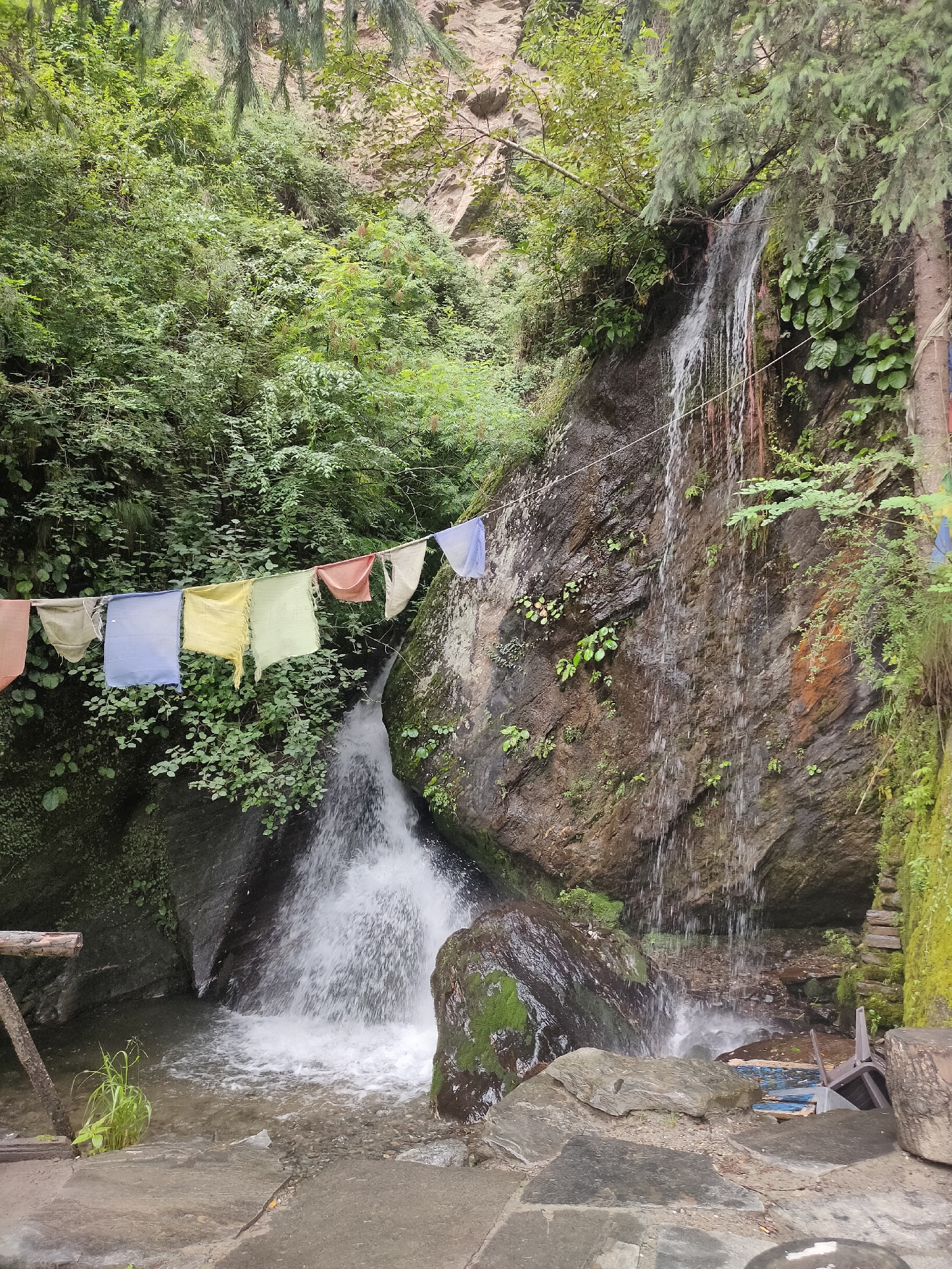 In the city of Jana, India, a waterfall cascades down a rocky cliff face. Prayer flags, in colors of blue, yellow, green, red, and white, are strung across the scene, fluttering in the breeze. Lush green foliage surrounds the waterfall and clings to the rocks, creating a vibrant and natural setting. The water crashes into a pool below, creating a misty spray. A large, moss-covered boulder sits in the foreground, with scattered rocks and a rough path leading to the water's edge. To the right of the waterfall, a discarded plastic chair rests against the rocks, adding a touch of human presence to the otherwise serene natural landscape.