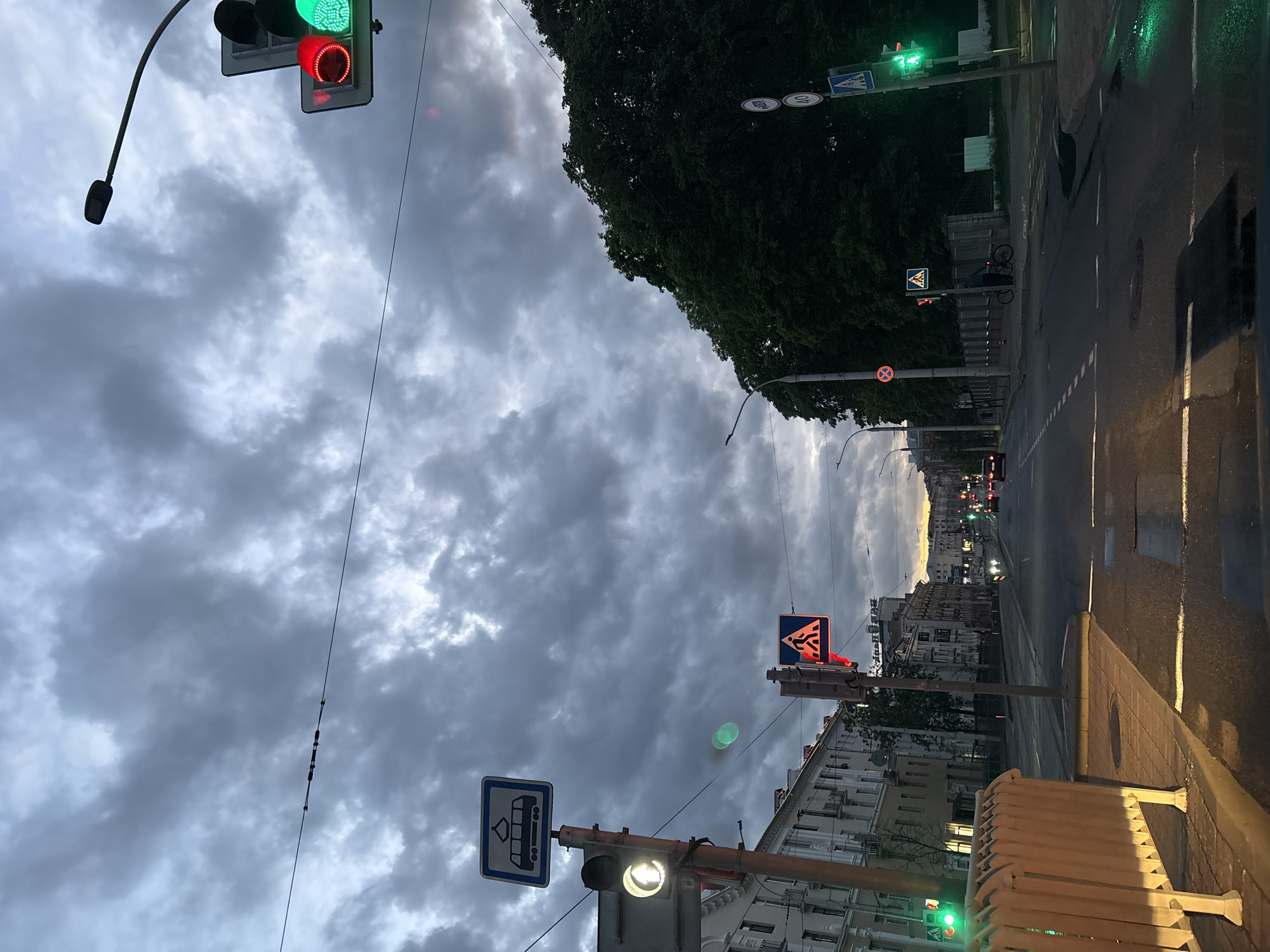 The image depicts a street in Minsk, Belarus, on a cloudy evening. The street is lined with buildings, and there are several traffic lights and street signs visible. The traffic light closest to the camera is showing a red light, while further down the street, other traffic lights show green. The sky is filled with heavy, gray clouds. There are no people visible in the image. The street appears wet, suggesting recent or ongoing rain. The location seems to be a residential area with buildings of varying heights and architectural styles. A yellow retractable barrier stands at the side of the road. There are overhead wires running across the sky.