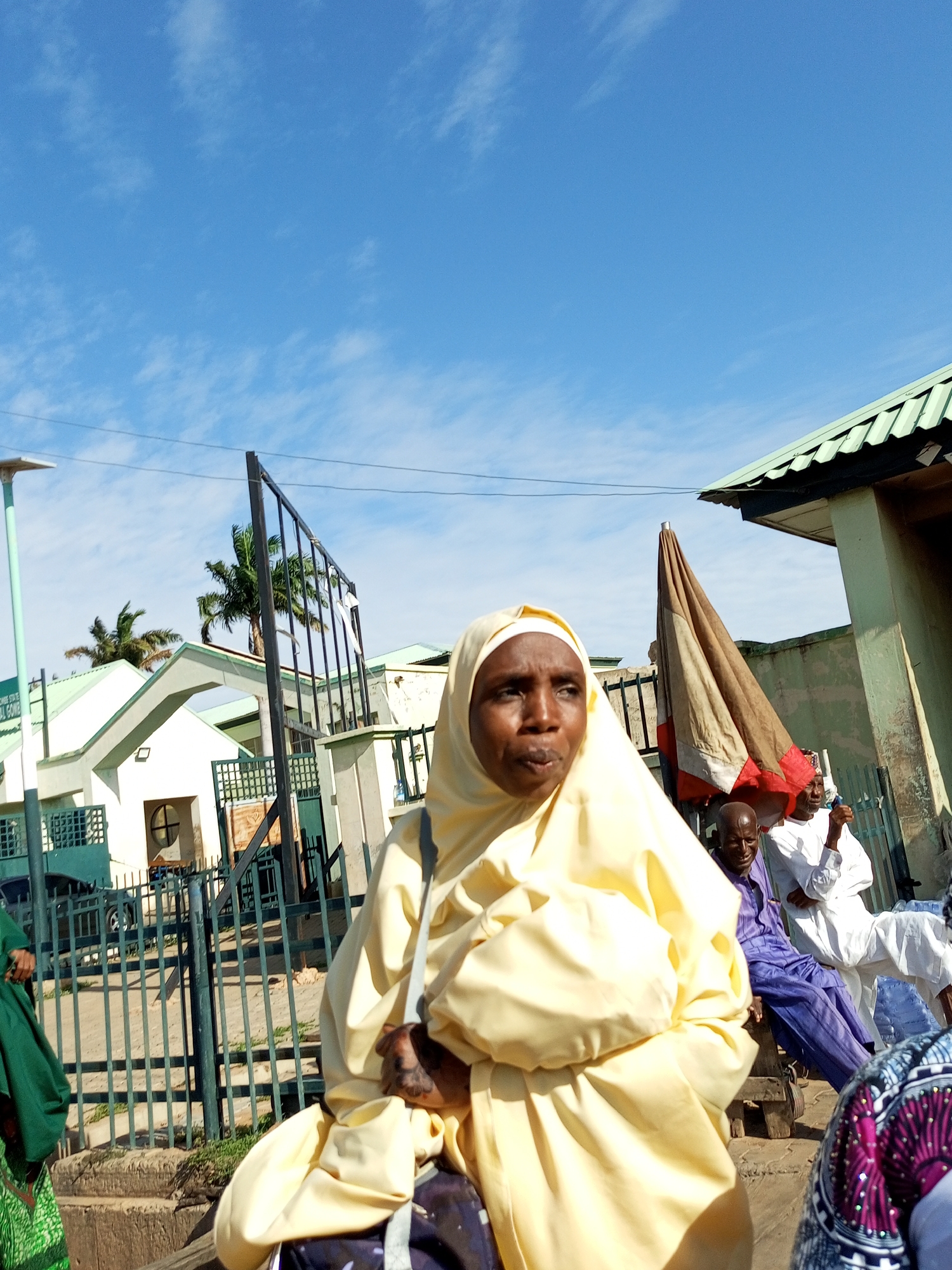 In Arawa, Nigeria, a group of people are gathered outdoors under a bright blue sky with wispy white clouds. The scene appears to be daytime, with the sun casting distinct shadows. The foreground is dominated by a woman wearing a pale yellow hijab and a coordinating garment. She is looking off to the side with a neutral expression. She carries a bag with a grey strap. Behind her, a large, partially furled umbrella with brown, white, and red panels stands in front of a building. Several other individuals are visible: a man in a purple striped outfit sits on a wooden bench, looking towards the umbrella. Further back, a man in a white garment sits to the right, his right arm raised. In the background, there is a white building with a green roof and a fence. The building has an arched entrance and a sign with green lettering, though the text is not fully legible. Palm trees are visible behind the building, suggesting a warm climate. A metal gate with vertical bars is also prominent. The overall impression is one of a casual gathering in a public outdoor space.