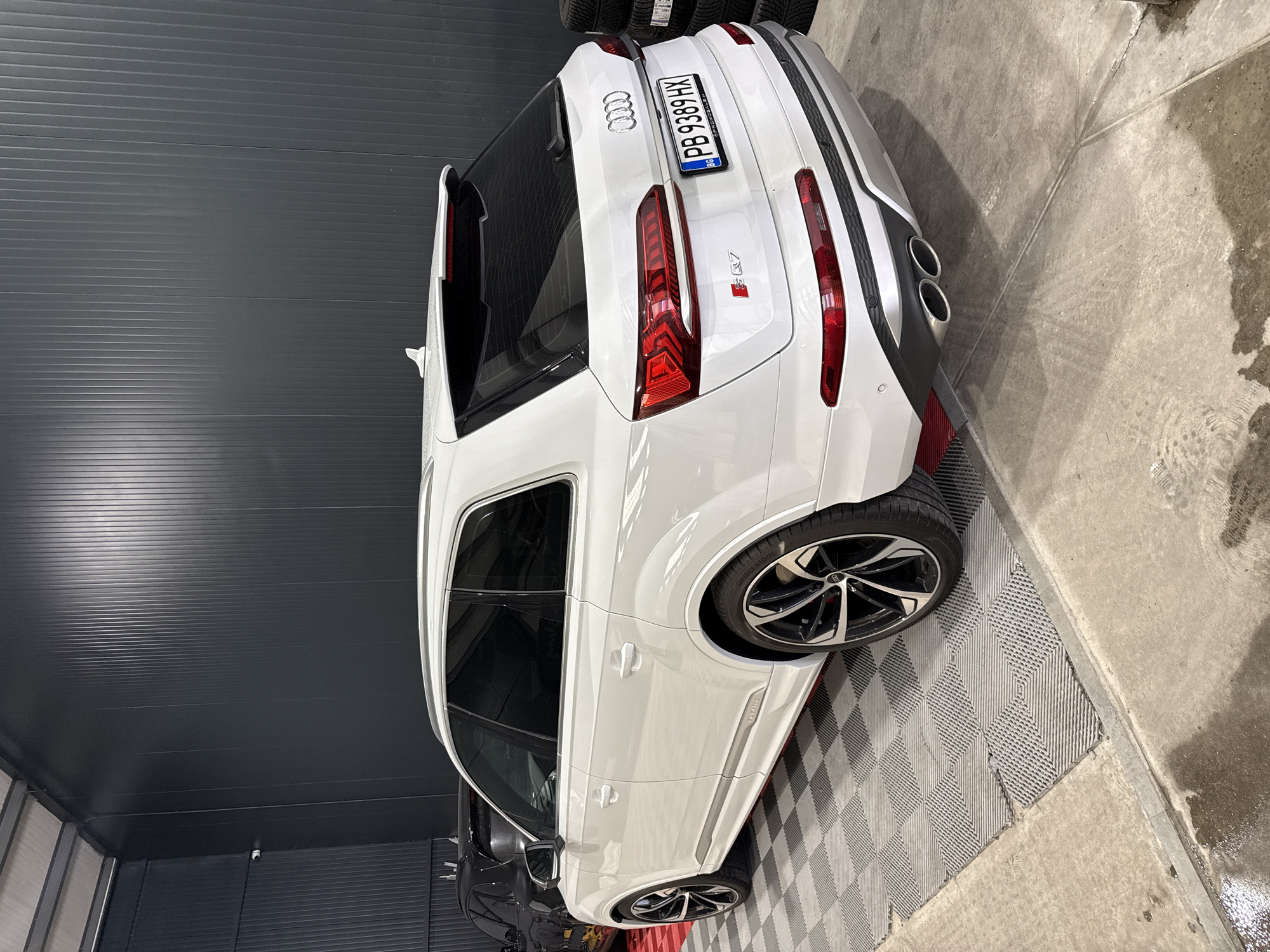 A white Audi SQ7 is parked inside a garage or car detailing facility in Plovdiv, Bulgaria. The license plate reads "PB 9389 HX". The car is angled slightly, showcasing its rear design with taillights, four exhaust pipes, and the Audi logo. The garage has a gray corrugated metal wall and a patterned gray floor. A black car is partially visible behind the SQ7. The floor area transitions from the patterned floor to a concrete surface with some damp patches. Stacks of tires are visible in the upper corner of the image.