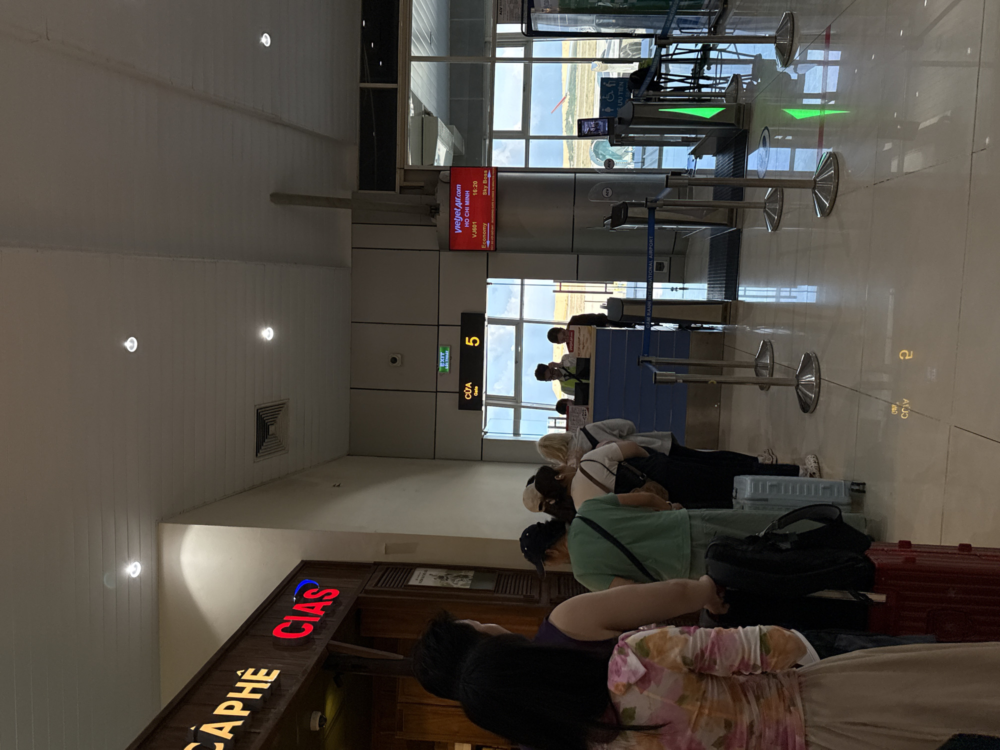 The image shows a departure gate at an airport, likely Cam Ranh International Airport in Bac Cam Ranh Ward, Vietnam, based on the sign that reads "RAM RANH INTERNATIONAL AIRPORT". A Vietjet Air flight, VJ601 to Ho Chi Minh City, is scheduled to depart at 16:20. Several passengers stand in line at Gate 5, presumably waiting to board. A cafe named "CAPHE CIAS" is nearby. The interior of the airport has a tiled floor, bright overhead lighting, and gate barriers. The outside visible through the glass windows features a runway.