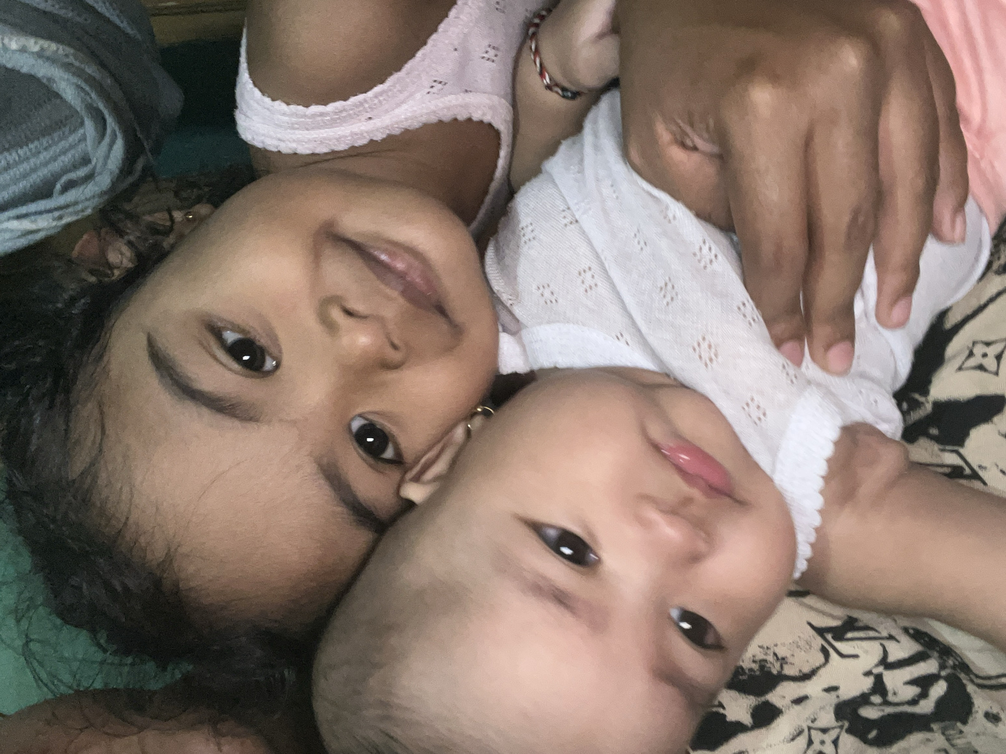 In Kruengmate, Indonesia, two young children are captured in a close-up, head-and-shoulders portrait. A toddler with dark, curly hair rests her head on the shoulder of a baby who is being held. The toddler gazes directly at the camera with wide, dark eyes and a slight smile. The baby, facing slightly away, also looks towards the viewer with large, expressive eyes. Both children are wearing light-colored clothing. The baby is dressed in a white garment with a delicate pattern, while the toddler wears a pink tank top. The background is softly blurred, with hints of green and gray fabric. The overall impression is one of warmth and closeness between the siblings.