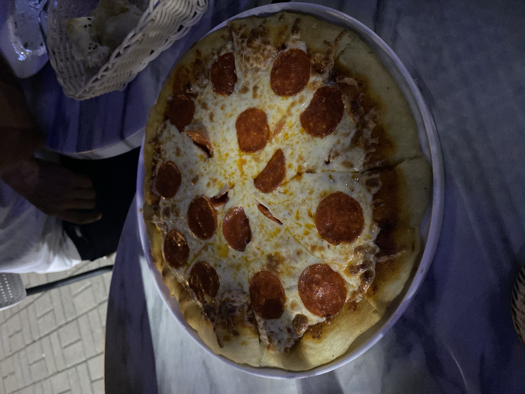 A pepperoni pizza sits on a white plate on a marble-topped table. The pizza is round with a golden-brown crust and is covered in melted cheese and pepperoni slices. In the background, a white basket filled with bread is visible. A person wearing a white shirt and dark pants is partially visible on the right side of the image. The scene appears to be outdoors in a city setting, possibly Bamboura, Abkhazia, as indicated by the tiled ground and outdoor furniture. The lighting suggests it is evening or nighttime.