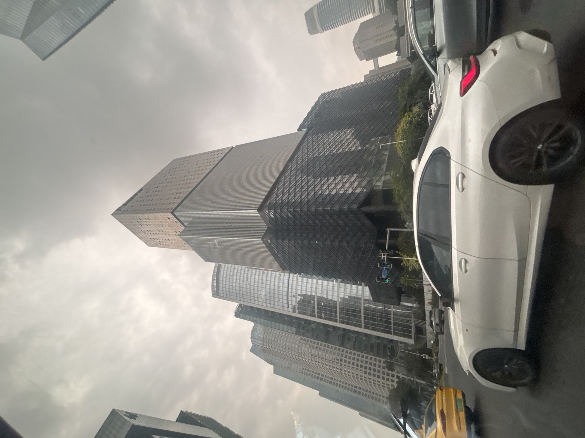 The image captures a street view in Tianhe District, China, dominated by a cluster of modern skyscrapers under an overcast sky. In the foreground, a white sedan is parked or moving on the wet road. To its left, a yellow taxi is visible. The backdrop features a complex of tall buildings with distinctive architectural styles, including glass facades and angular designs. One particularly large building in the center has a dark, textured lower section and a lighter, segmented upper section. Traffic lights are visible on the right side of the road, indicating an intersection. The overall scene suggests a bustling urban environment, possibly during or after rainfall.