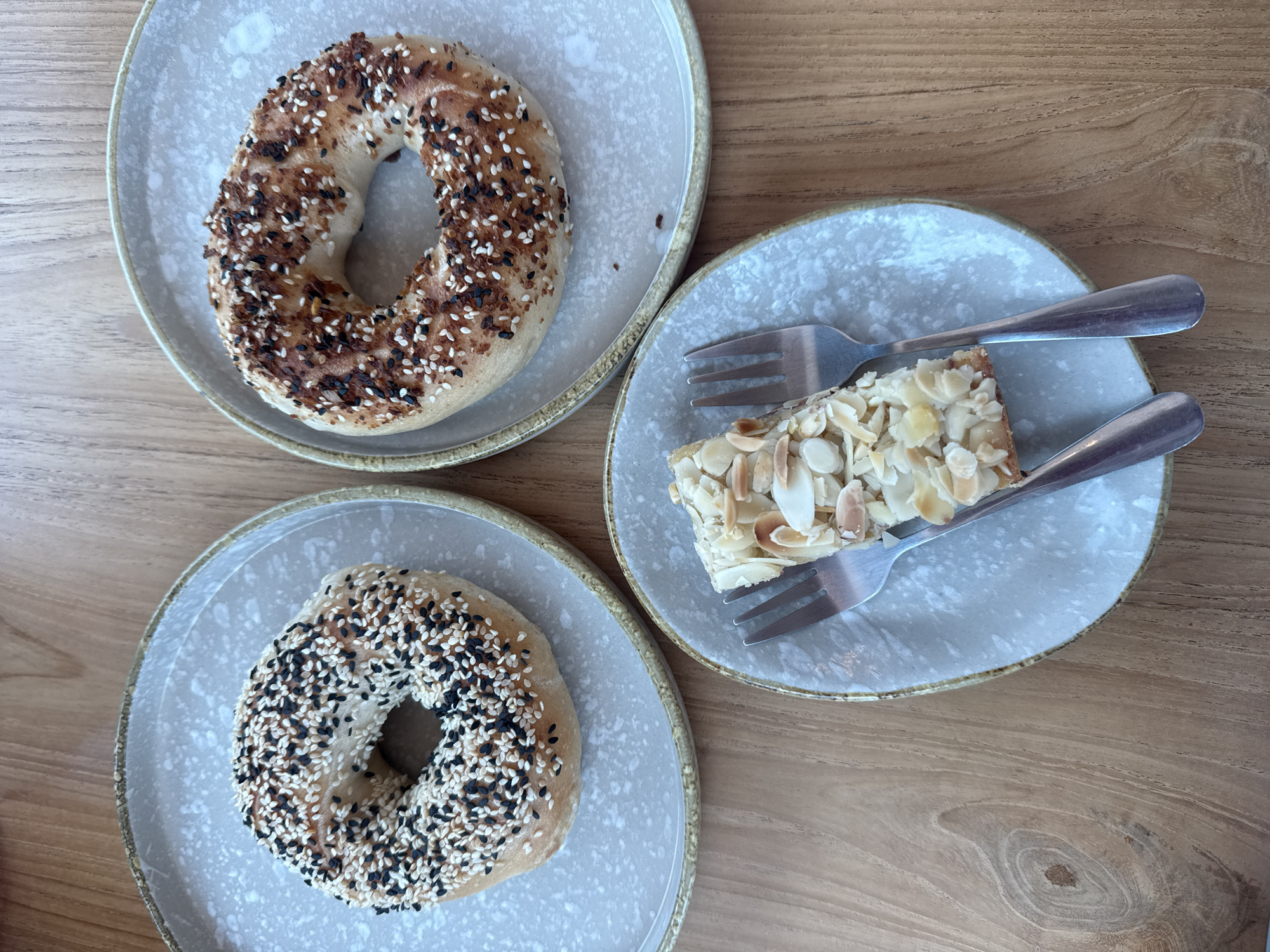 Two bagels, one with black and white sesame seeds and the other with a mixture of seeds including poppy and what appear to be brown spices, are presented on separate, speckled gray plates. A slice of cake, topped with sliced almonds and possibly sugar, sits on a third, similar speckled gray plate, accompanied by two silver forks. All items are arranged on a light brown wooden surface with a visible wood grain pattern. The image is captured from a top-down perspective. The scene is not set in a specific city or district of Ko Samui, Thailand; it appears to be a generic food presentation. There are no people or interactions depicted in the image.
