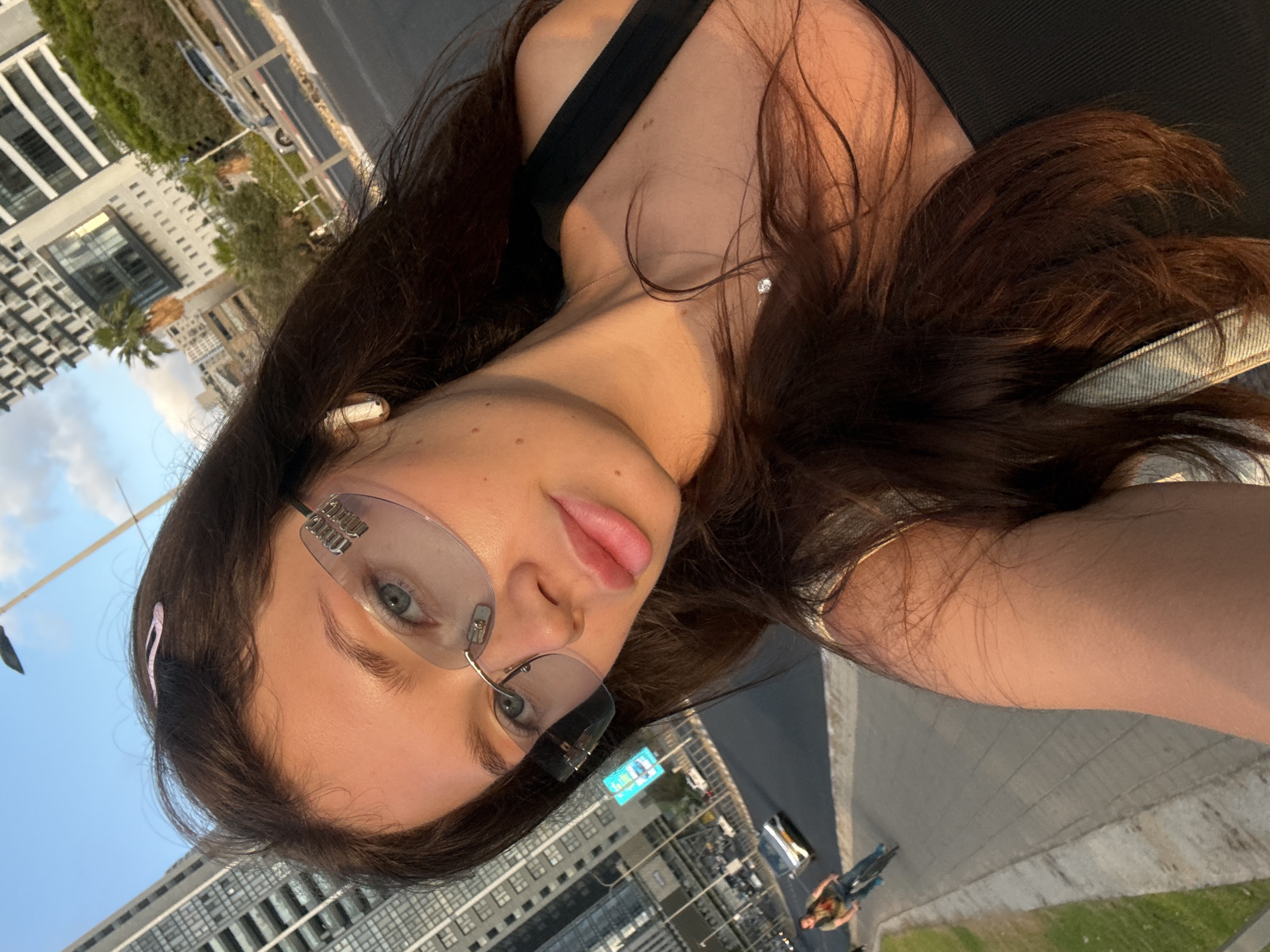 A young woman poses for a selfie in Tel Aviv, Israel. She is wearing rimless glasses, an earbud in her left ear, and a black top with a thin strap. She also has a silver necklace with a diamond pendant. Her hair is long and brown, with a pink hair clip on the left. Buildings are visible in the background, along with city streets, cars, and a person riding a bicycle.