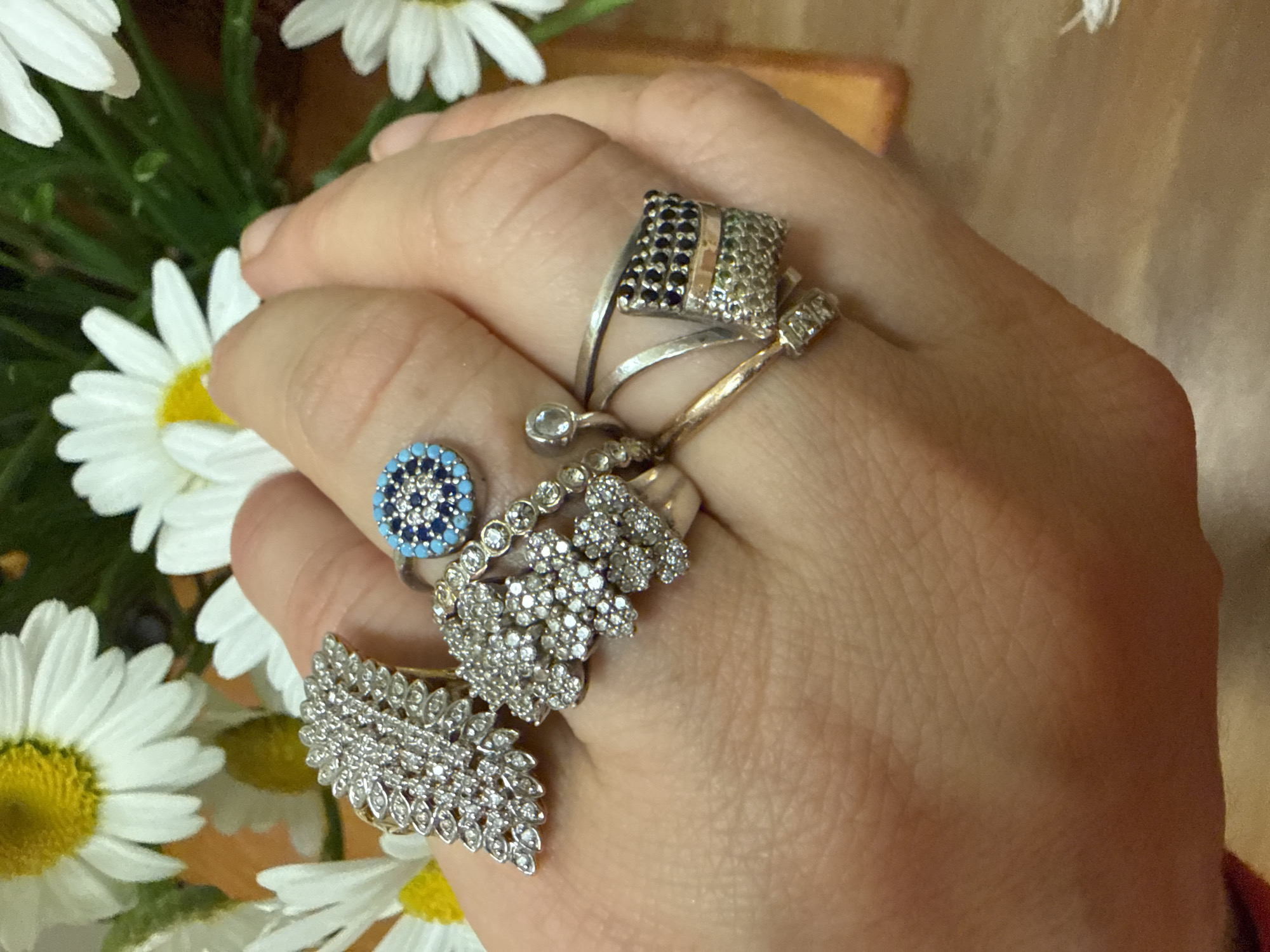 A close-up, overhead shot shows a person's hand adorned with several rings, with daisies in the background. The hand is positioned on a wooden surface. The rings are made of silver and adorned with various gemstones, including blue and clear stones, and some with intricate patterns. The daisies are white with yellow centers, and some green foliage is visible behind them. The lighting is natural and casts soft shadows. The overall scene is casual and domestic. There is no indication of the location being in Rome, United States of America, or any specific city.
