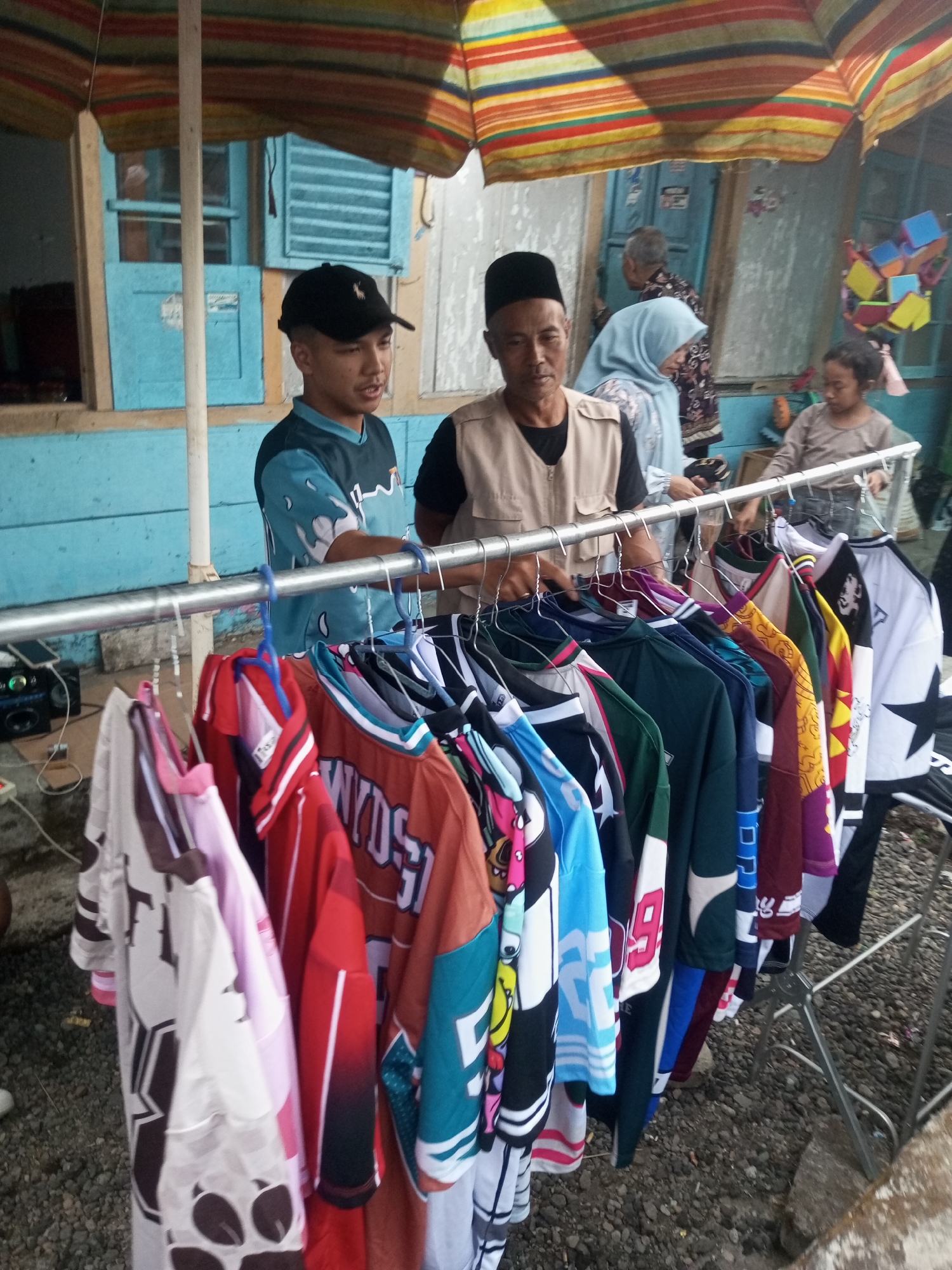 In Pandansari, Indonesia, a roadside stall displays a variety of shirts on hangers. Two men stand near the stall, one wearing a black cap and a teal and black patterned shirt, the other a tan vest over a black shirt and a black cap. Behind them, a woman in a light blue headscarf and another person are visible. The stall is set up under a colorful striped umbrella, with a metal rack holding the clothing. The ground is covered with gravel. In the background, a building with blue walls and windows can be seen.