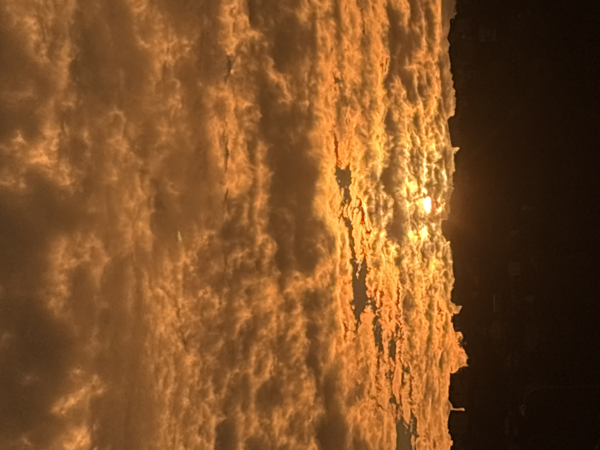 The image depicts a dramatic sunset over a silhouetted cityscape. The sky is dominated by a dense layer of textured, orange and brown clouds, illuminated from within by the setting sun. The sun itself is a bright, small orb peeking through a gap in the clouds near the center of the frame. The clouds are detailed and appear to be low-lying, creating a textured ceiling. Below the sky, a dark, silhouetted ridge line represents the urban landscape, with the indistinct shapes of buildings and possibly trees visible. The overall impression is one of intense color and atmospheric drama. The image does not provide any specific visual cues to definitively place it in Sochi, Russia; it is a generic depiction of a sunset over a city. There are no people, specific actions, or interactions visible. The environment is clearly an outdoor scene at dusk.
