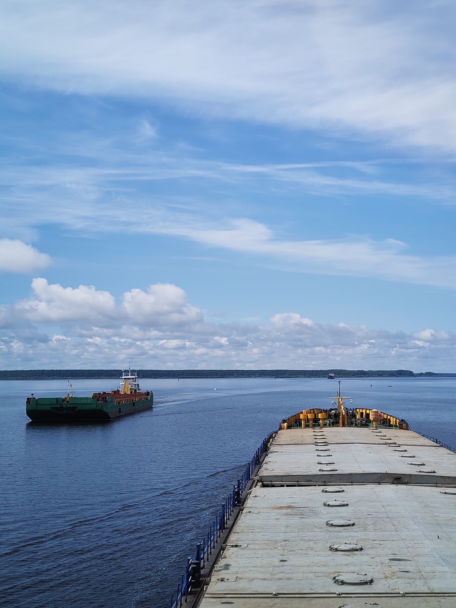 The image shows a wide, calm body of water, likely a river or lake, under a partly cloudy blue sky. Two barges are visible. In the foreground, the deck of a large barge, characterized by its flat, gray surface with numerous circular hatches, stretches towards the viewer. The barge has a railing with blue posts. In the middle distance, a smaller green barge with a white cabin structure is moving away from the viewer. The background features a distant, tree-lined shore. The scene is consistent with views of the Rybinsk Reservoir near Cherepovets, Russia, specifically on the Sheksna River, where such barge traffic is common. There are no people visible in the image. The overall impression is one of calm maritime activity on a bright day.