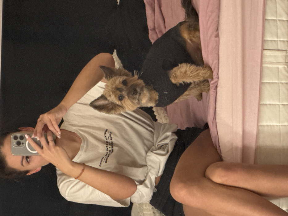 A woman takes a selfie with her iPhone in a mirror. She is wearing a white t-shirt and black shorts. A small dog is wearing a black sweater and lying to her right on a pink blanket. A white quilted bed is visible on the right side of the frame, while a black surface can be seen on the left side of the frame.
