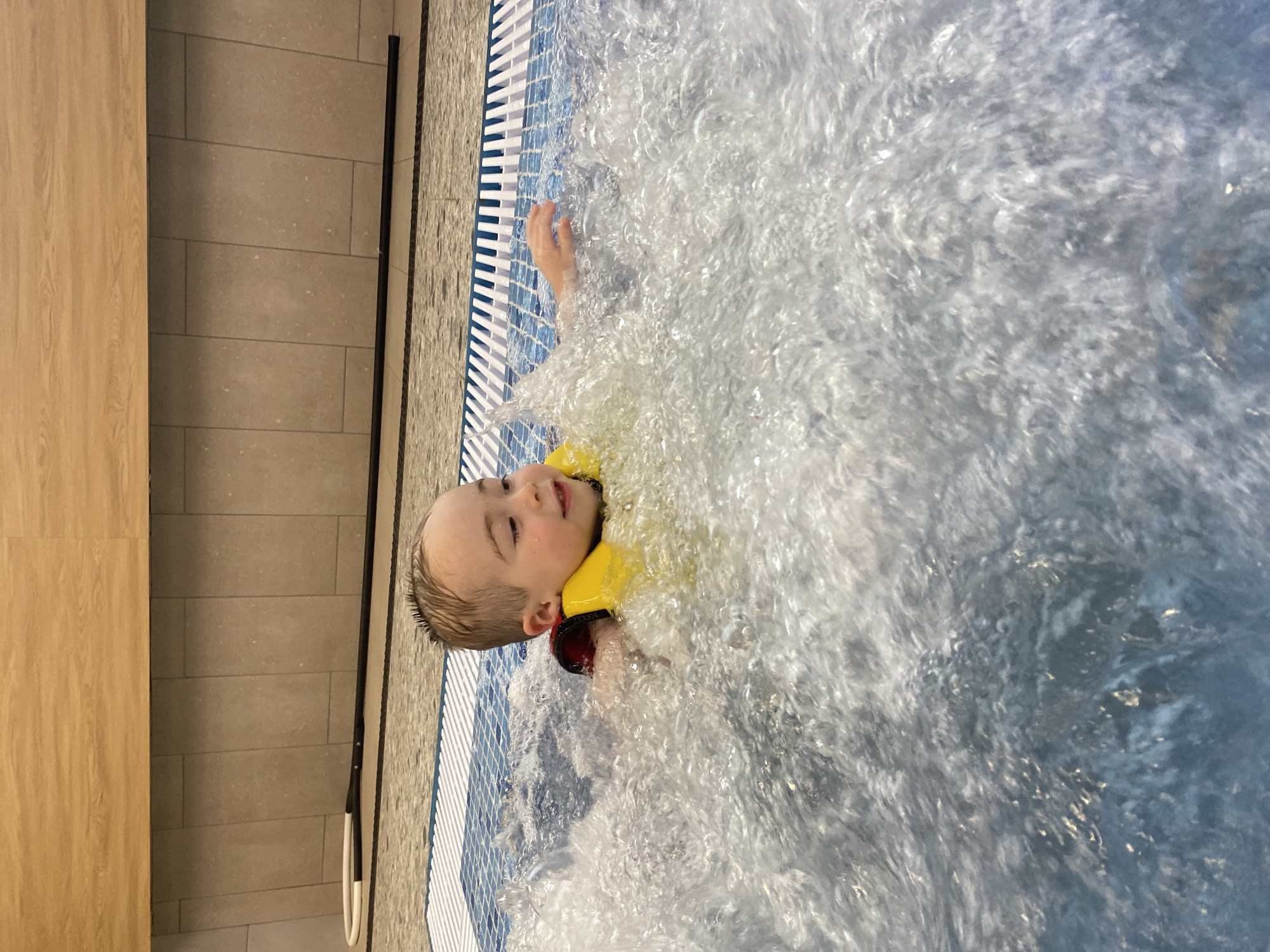 A young child, wearing a yellow buoyancy aid over a red garment, is partially submerged in a heavily aerated body of water, indicative of a jacuzzi or whirlpool feature. The child has short, light-colored hair and a discernible smile, gazing towards the right side of the frame. The water around the child is actively bubbling and turbulent. Behind the child, a pool edge with a blue and white mosaic tile pattern is visible. Beyond this edge, a vertical wall surfaced with large, light grey rectangular tiles extends upwards, topped by a horizontal section of light wood-grained paneling. A black pole, featuring a white and black circular loop at its end, is positioned on the tiled edge to the left. The scene is set indoors within an aquatic facility in the city of Votkinsk, Russia.