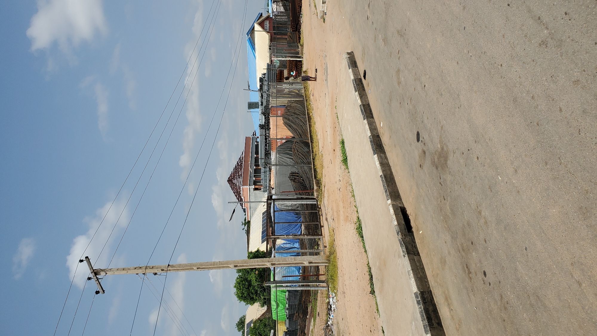 The image depicts a street scene in Jalingo, Nigeria. A concrete sidewalk with a drainage grate runs along the right side of the image, bordering a dirt lot. The lot is filled with construction materials, including stacks of rebar, tarps covering bundles, and what appear to be shipping containers. A single person is standing in the dirt lot, facing away from the camera. In the background, buildings with various roof types are visible, along with a telephone pole and overhead wires. The sky is blue with scattered clouds.