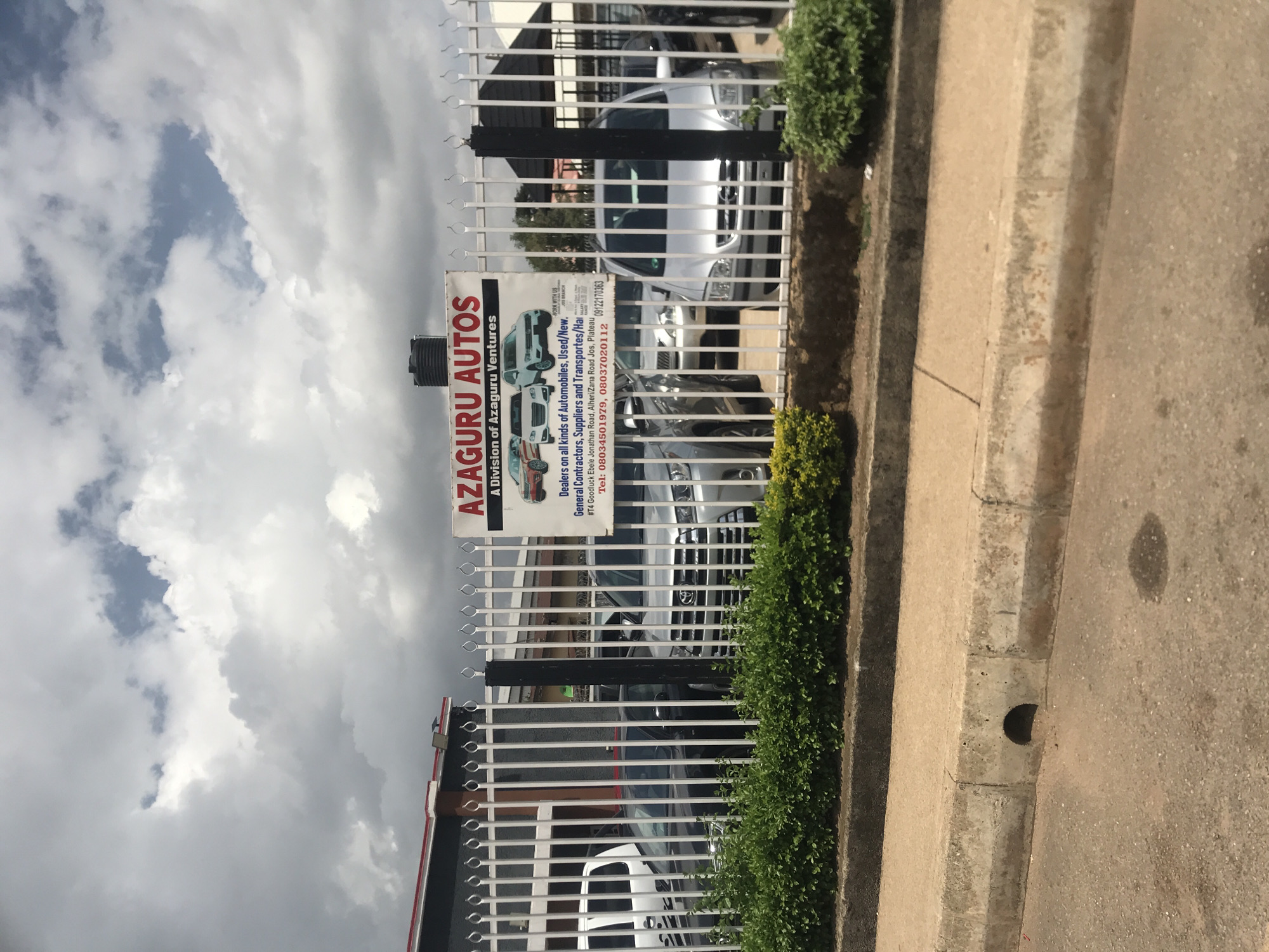 A white metal fence with vertical bars and decorative tops encloses a car lot. A sign on the fence reads "AZAGURU AUTOS A Division of Azaguru Ventures" and features images of cars. Below the main text, it states "Dealers on all kinds of Automobiles, Used/New. Contractors, Suppliers and Transportes/Ha". Further details include an address and phone numbers. Several cars are visible behind the fence, including a silver SUV, a black sedan, a white van, and other vehicles. Green hedges line the bottom of the fence. In the foreground, a concrete curb and asphalt road are visible, with a drain opening in the curb. The sky above is cloudy.